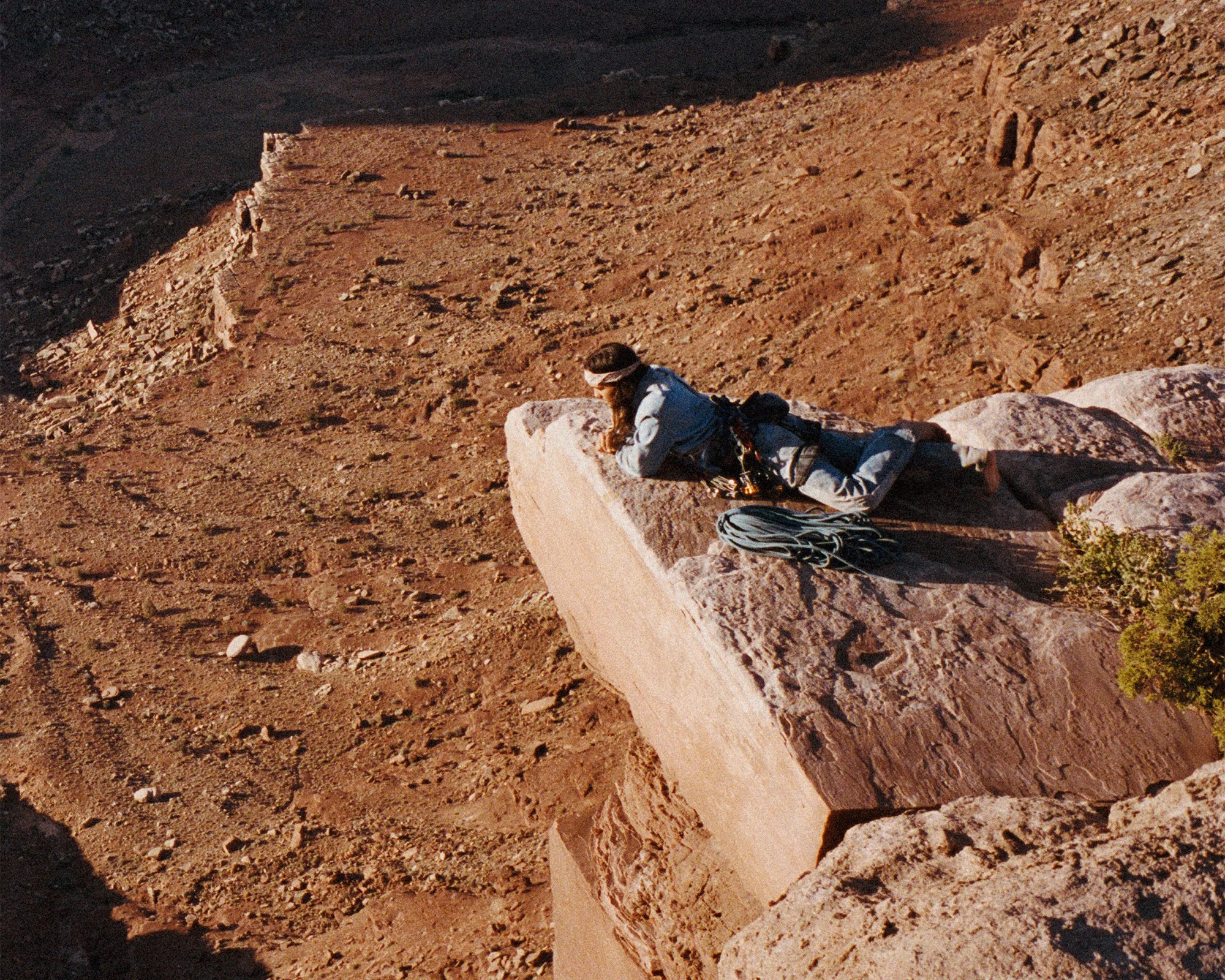 Person in blue climbing gear lying on a large rock ledge with coiled rope beside them in a desert landscape.