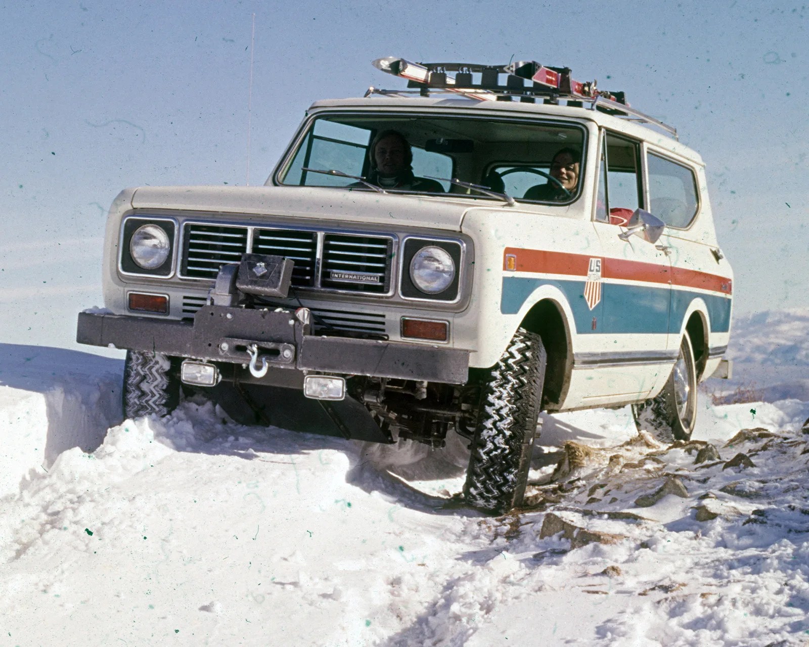 White International Harvester Scout SUV with red and blue stripes driving on snow-covered rocky terrain.