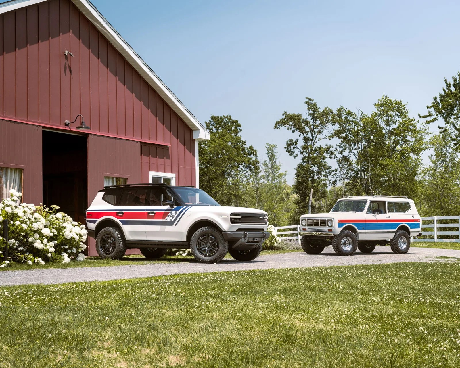 Two white SUVs with red and blue racing stripes parked near a red barn and white fence on a sunny day.