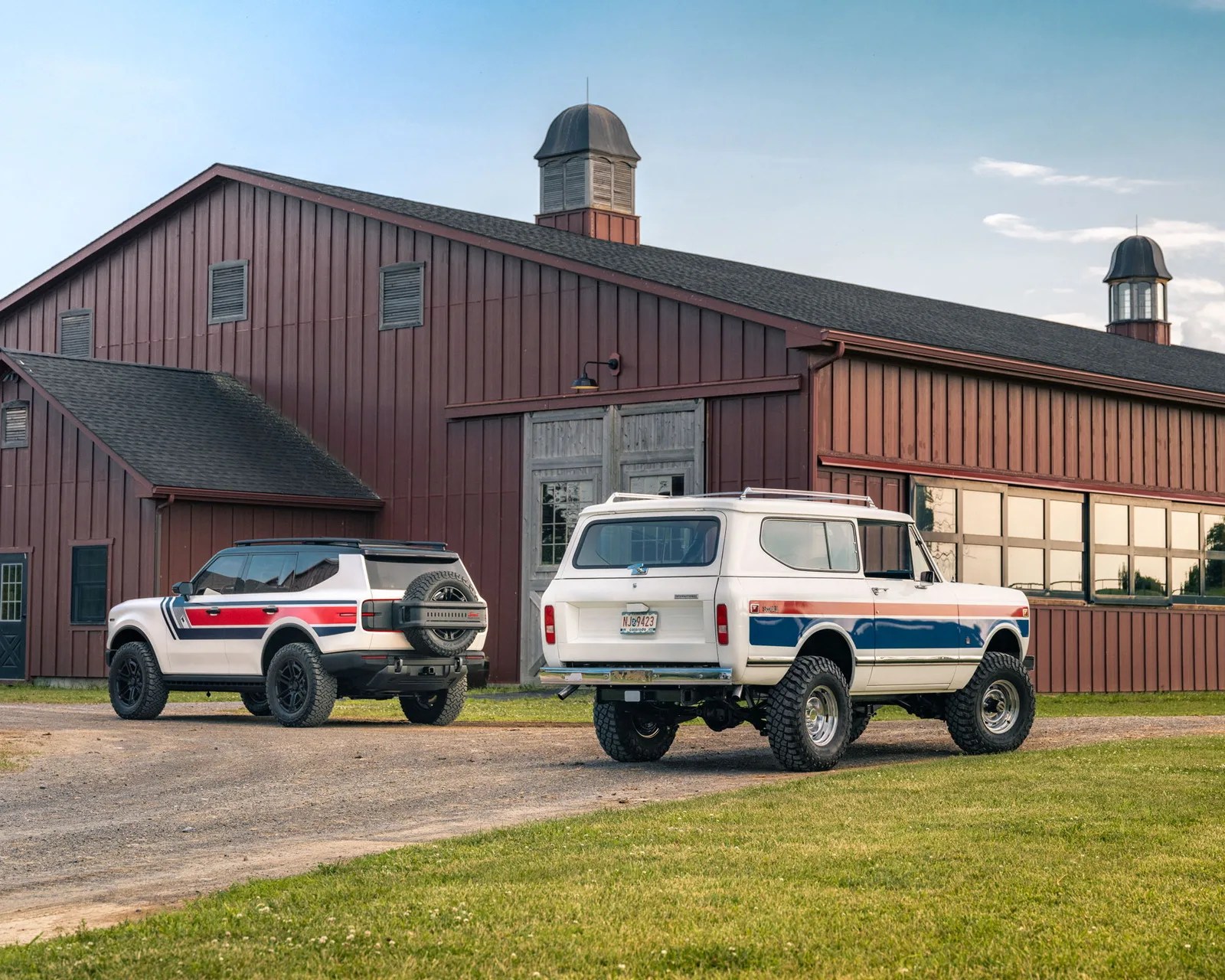 Two white SUVs with red and blue stripes parked on a gravel driveway in front of a large red barn.