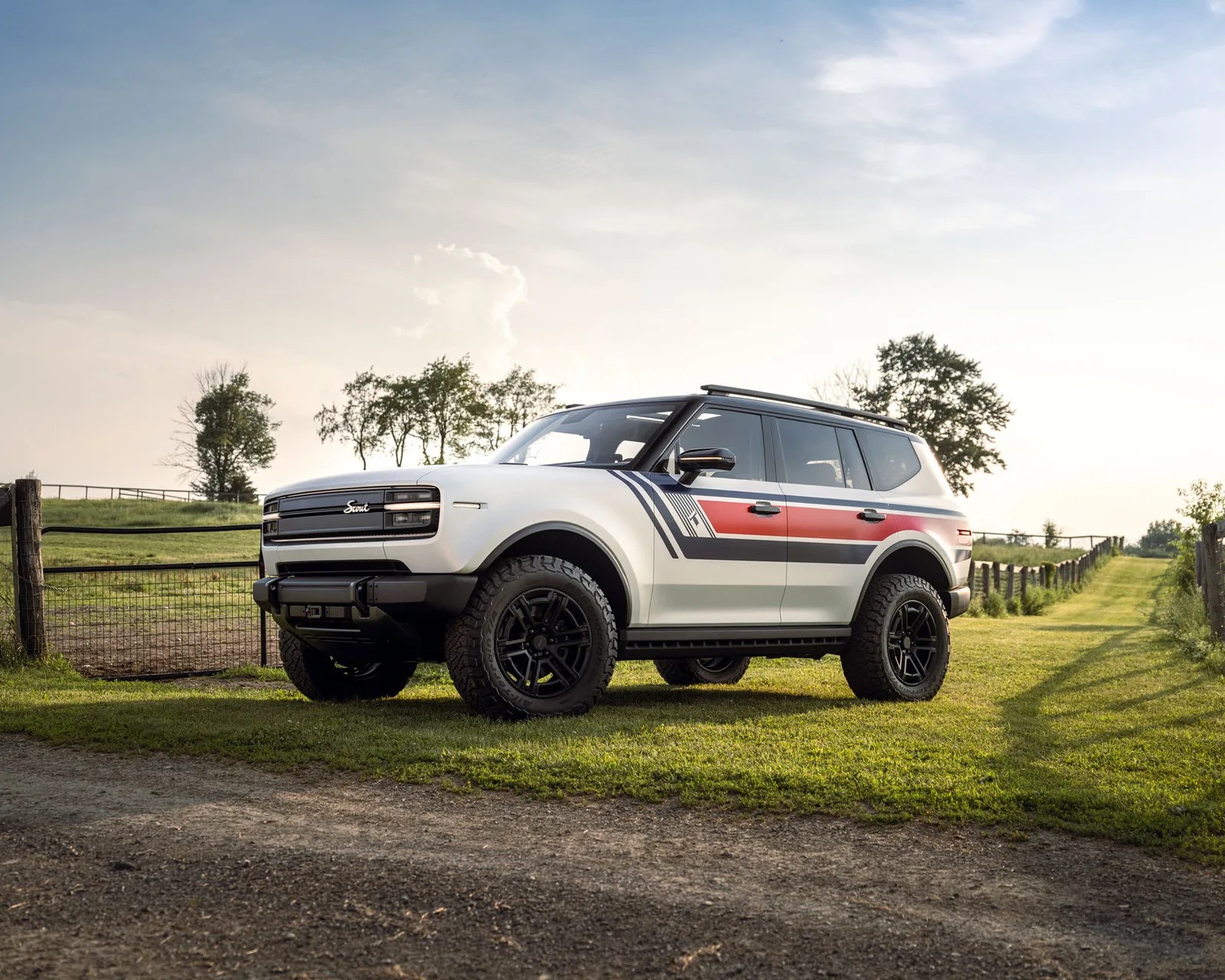 White SUV with black roof and red and black side stripes parked on grass near a dirt path and fence.