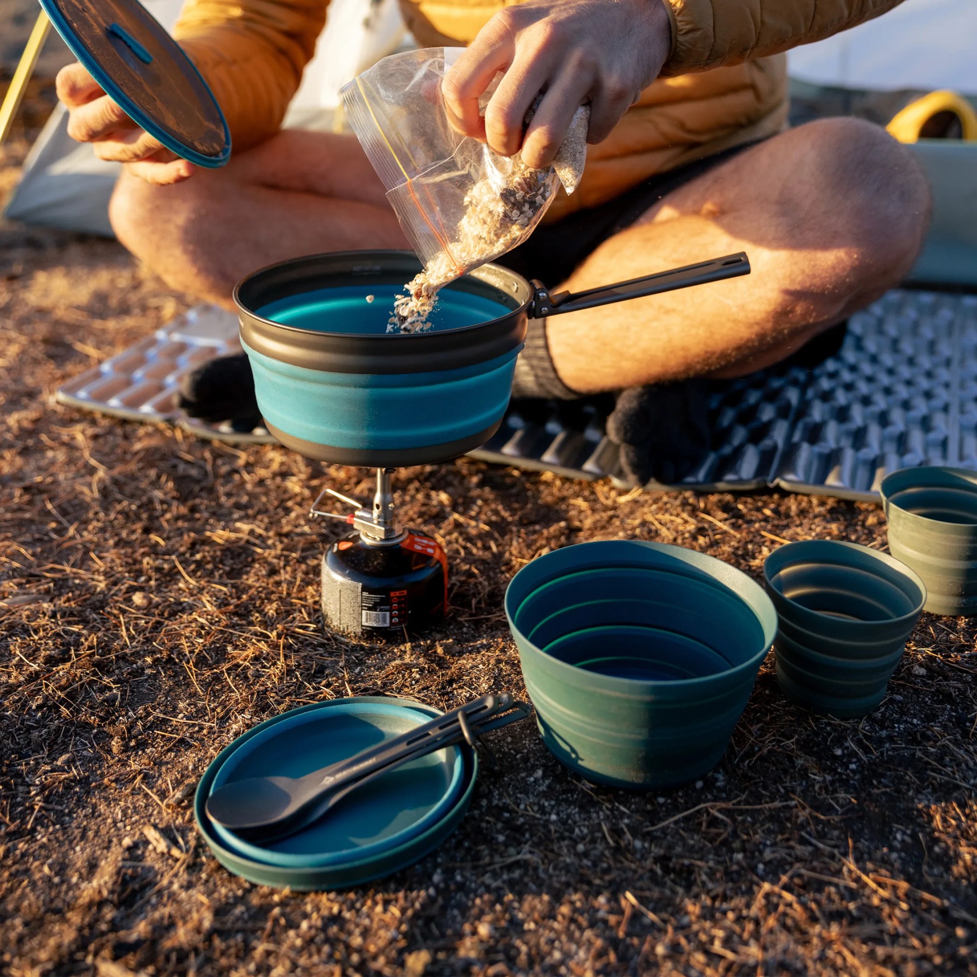 Person pouring dry oats from a plastic bag into a blue collapsible camping pot on a portable stove outdoors.