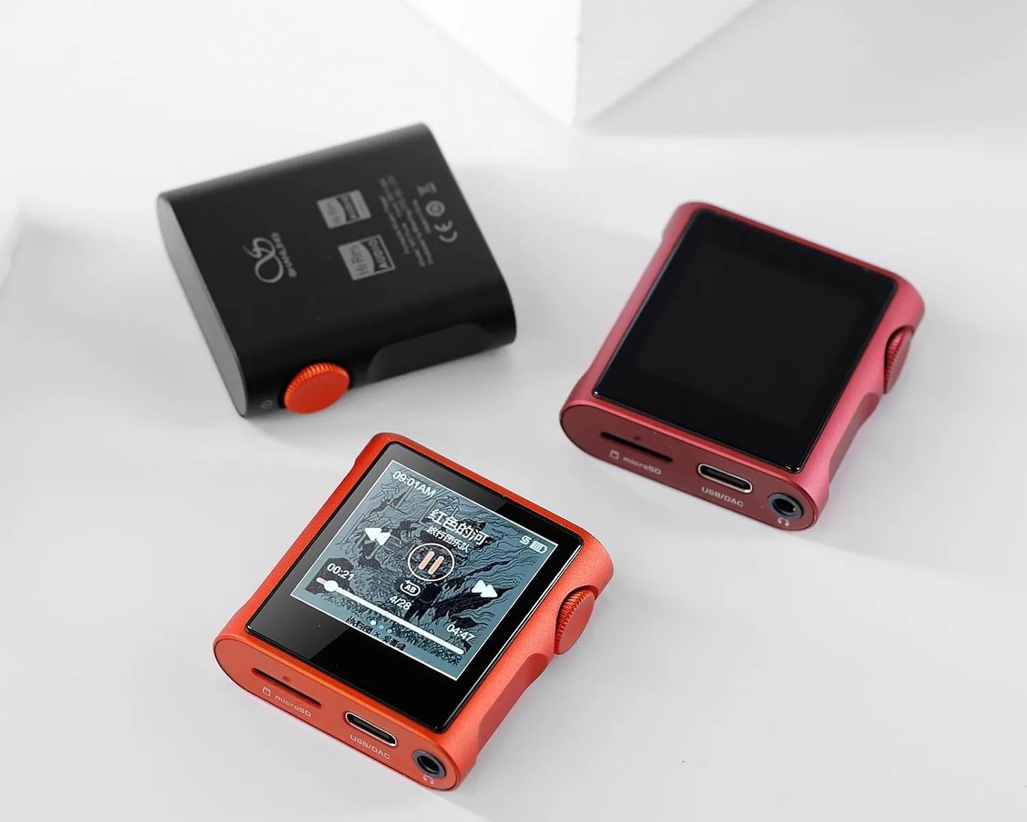 Three compact digital music players in black, red, and orange with screens and control knobs on a white surface.