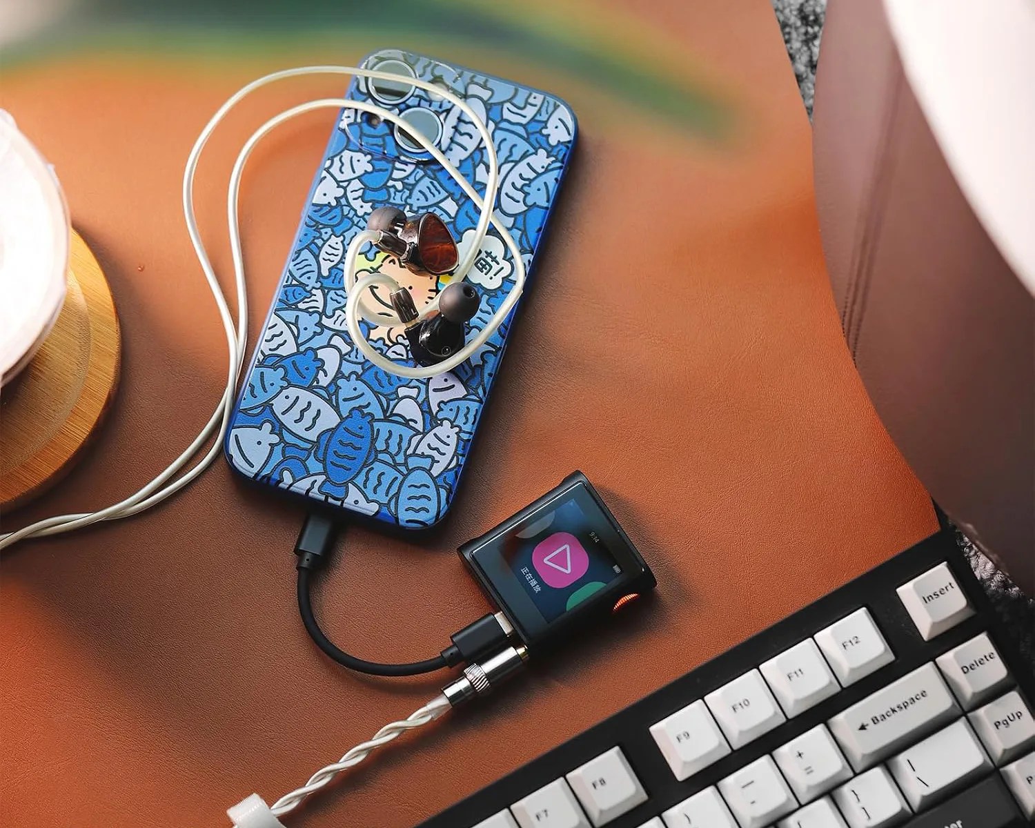Blue smartphone with a fish-patterned case, wired earphones, and a small black music player connected by cables on a brown surface next to a white keyboard.