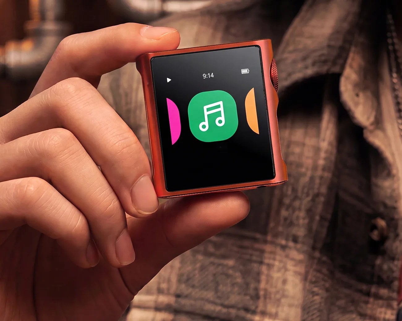 Hand holding a small square red digital music player with a touchscreen showing a green music note icon.