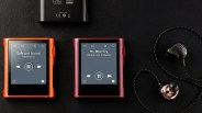 Two portable music players, one orange showing "Safe and Sound" by Capital Cities and one red showing "Mr. Blue Sky" by Electric Light Orchestra, next to black and copper wired earphones on a black surface.