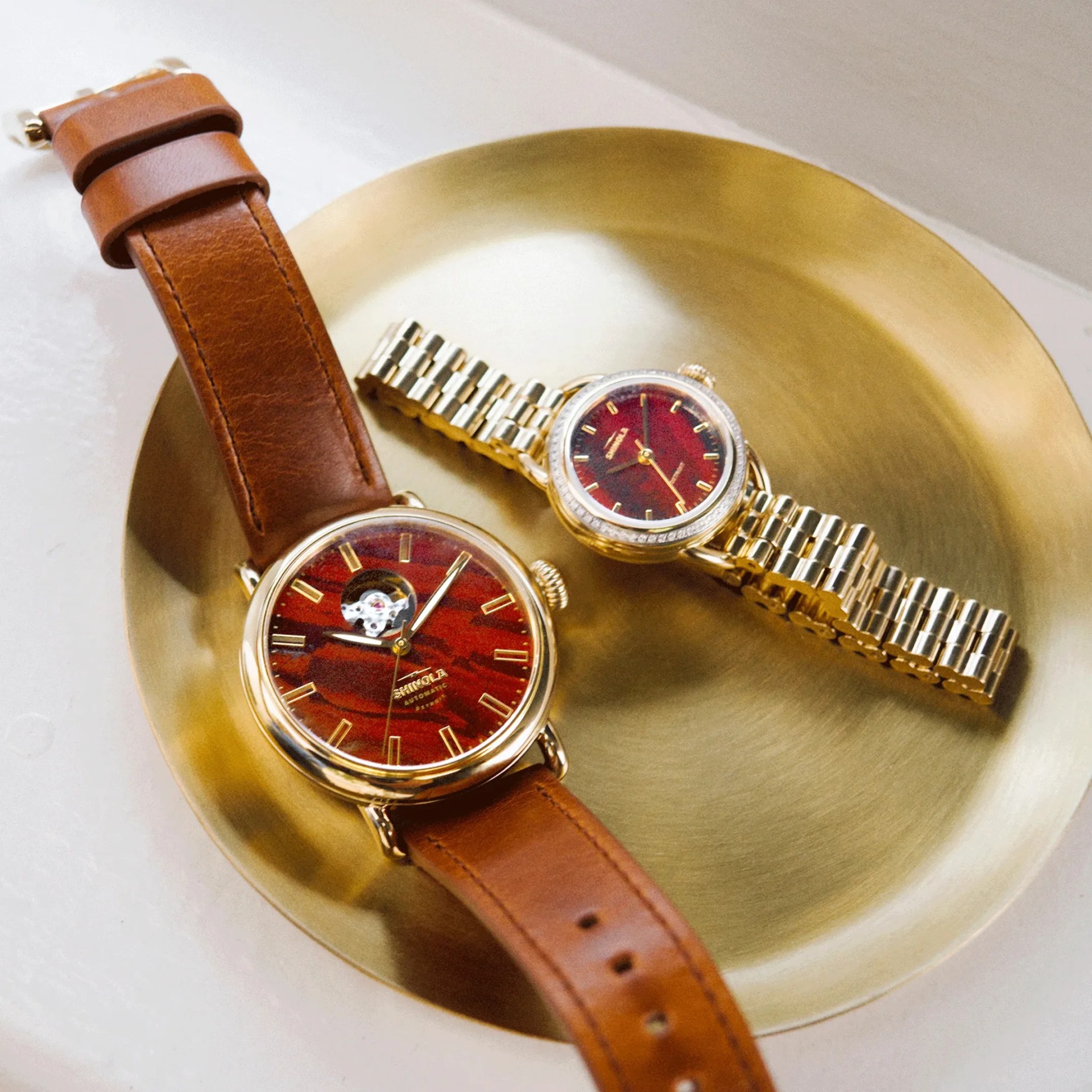 Two wristwatches with red dials and gold accents, one with a brown leather strap and the other with a gold metal bracelet, on a brass dish.