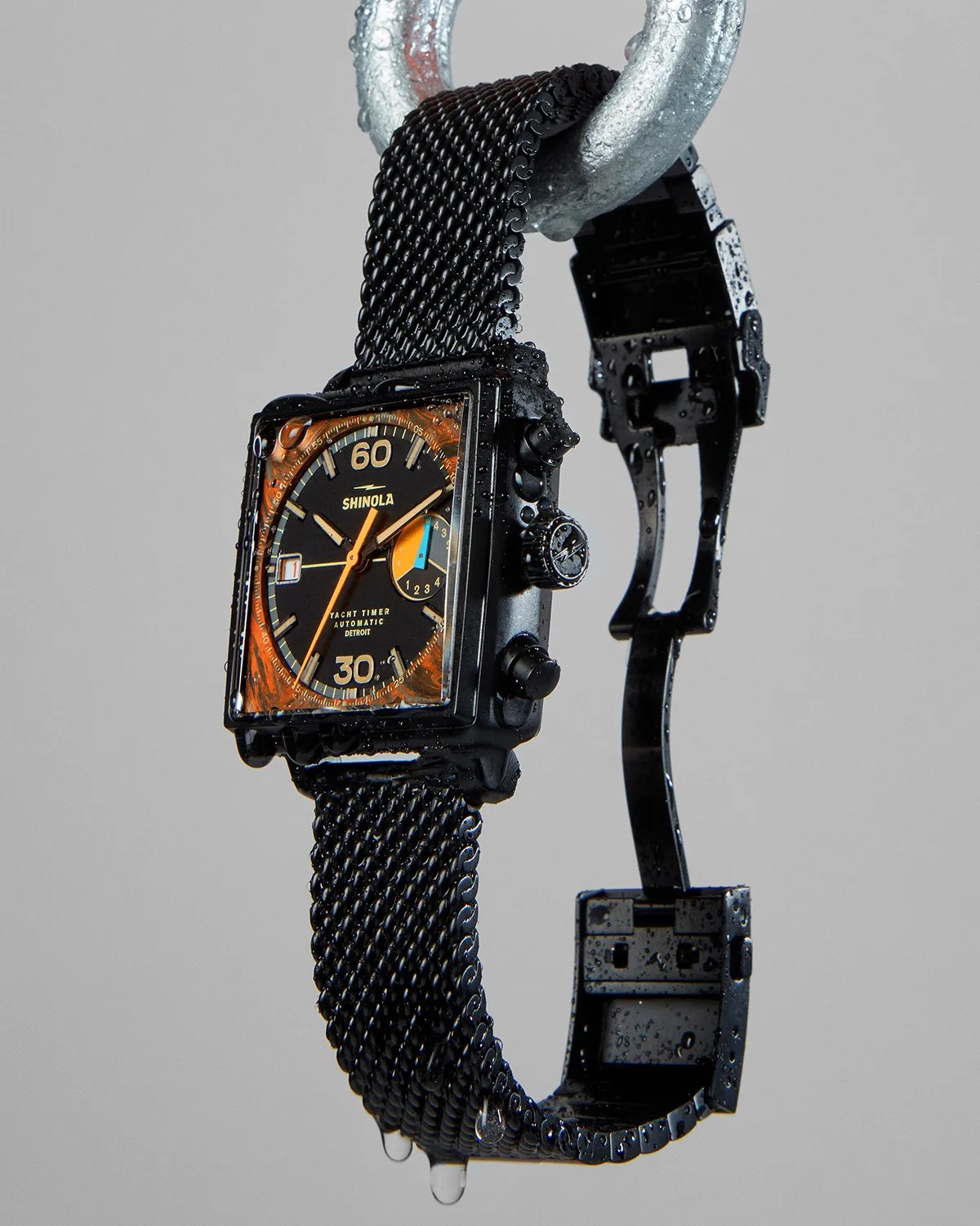 Black Shinola Yacht Timer watch with square face, orange accents, and black mesh strap hanging on a metal hook with water droplets.