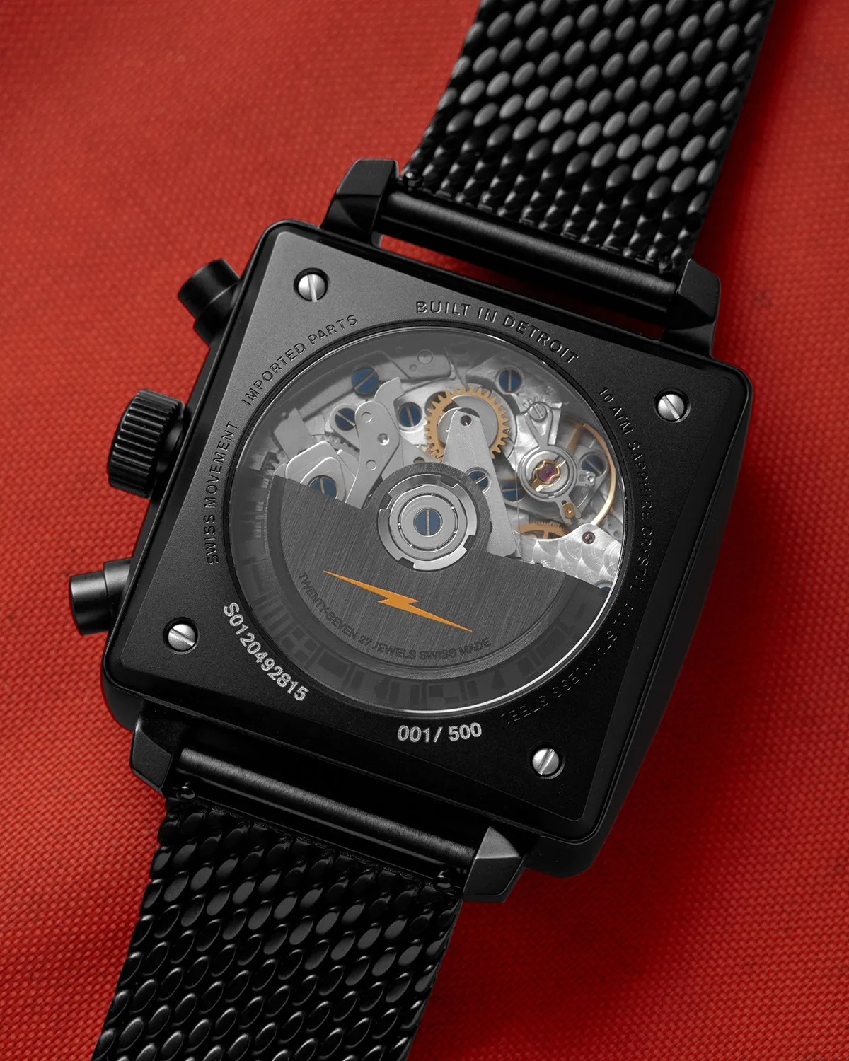 Black square watch with visible mechanical movement and black textured strap on red fabric background.