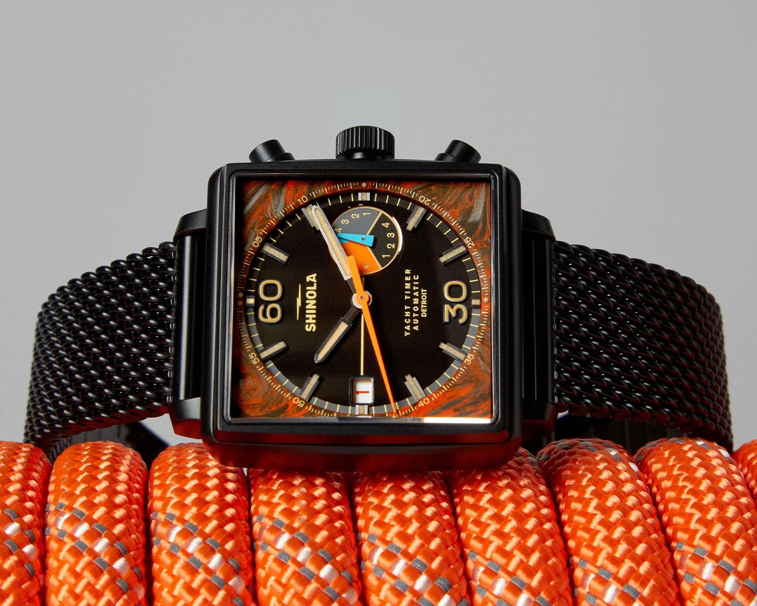 Square black Shinola Yacht Timer watch with orange and blue accents on a black mesh strap, resting on orange rope.