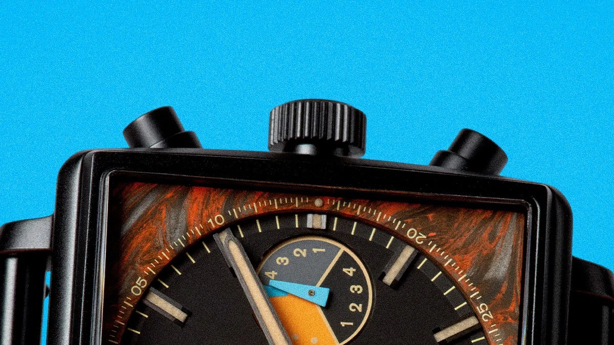 Close-up of a black square watch face with orange and blue accents and two black push buttons on a bright blue background.