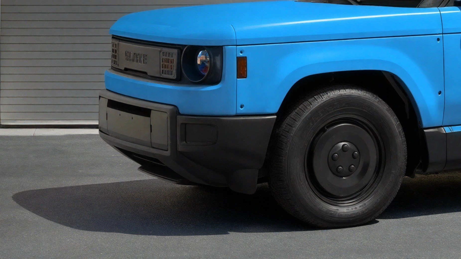 Front side of a blue electric vehicle with black bumper and wheel, parked on asphalt in front of a closed garage door.