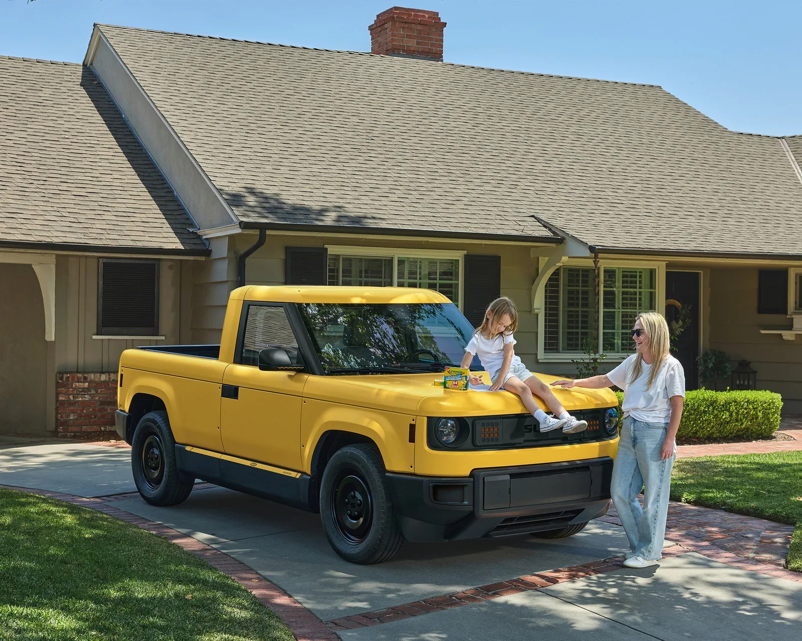 Yellow electric pickup truck parked in a driveway with a child sitting on the hood and an adult standing nearby.