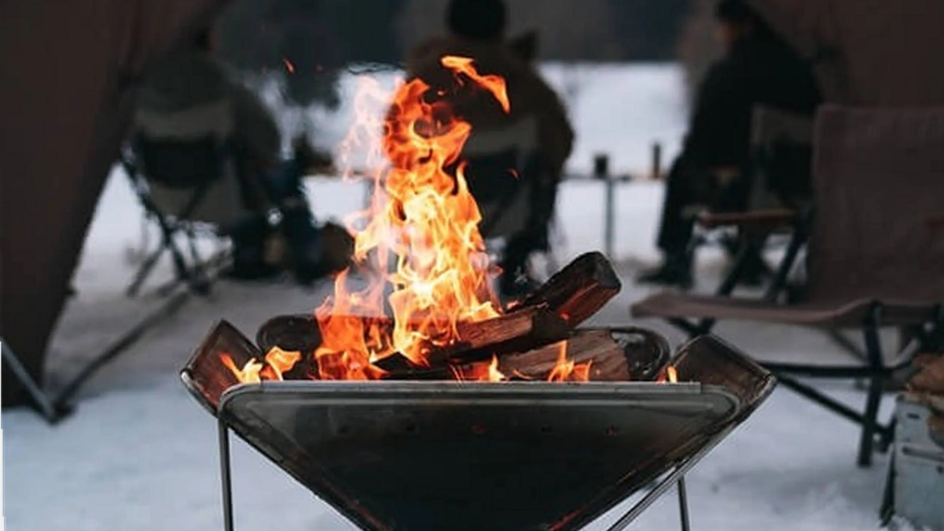 Metal fire pit with burning logs and orange flames in a snowy outdoor setting with chairs in the background.
