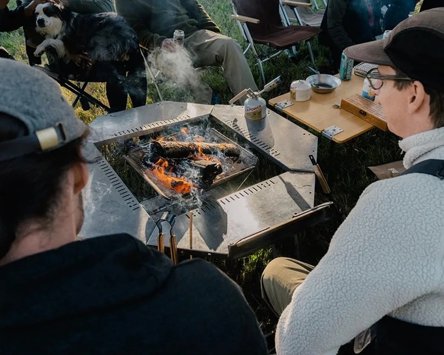 Outdoor metal fire pit with burning logs surrounded by people sitting on camping chairs and a dog on a lap.
