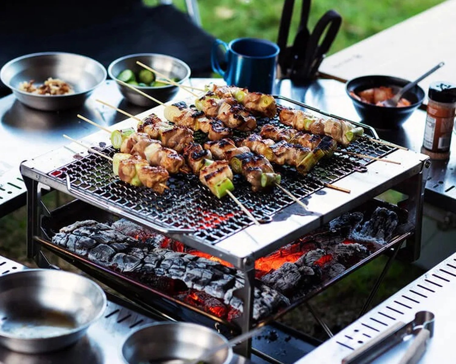 Charcoal grill with skewers of grilled meat and vegetables cooking over glowing coals on a metal table.