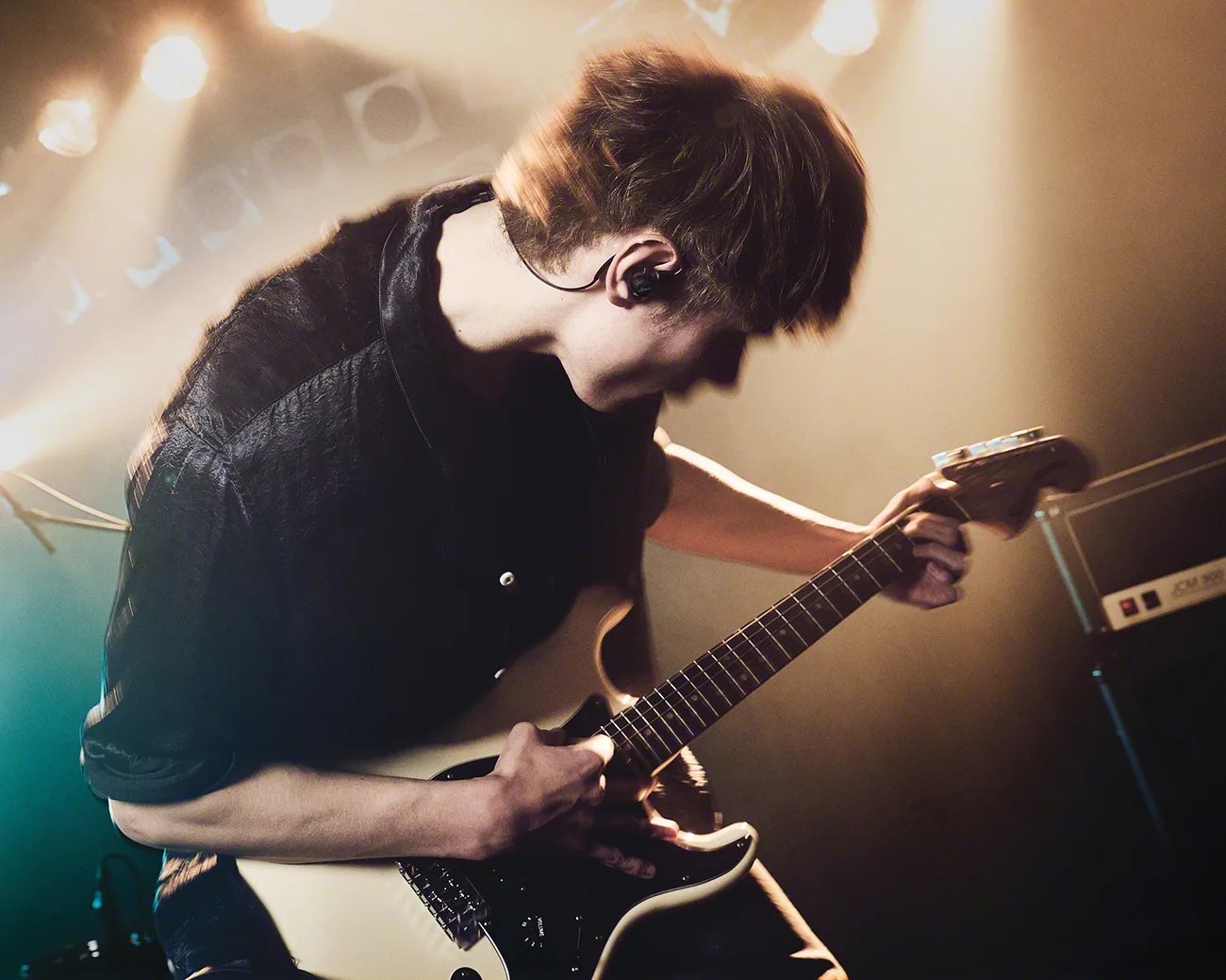 Person playing a white electric guitar on stage with warm lighting and wearing a black shirt.