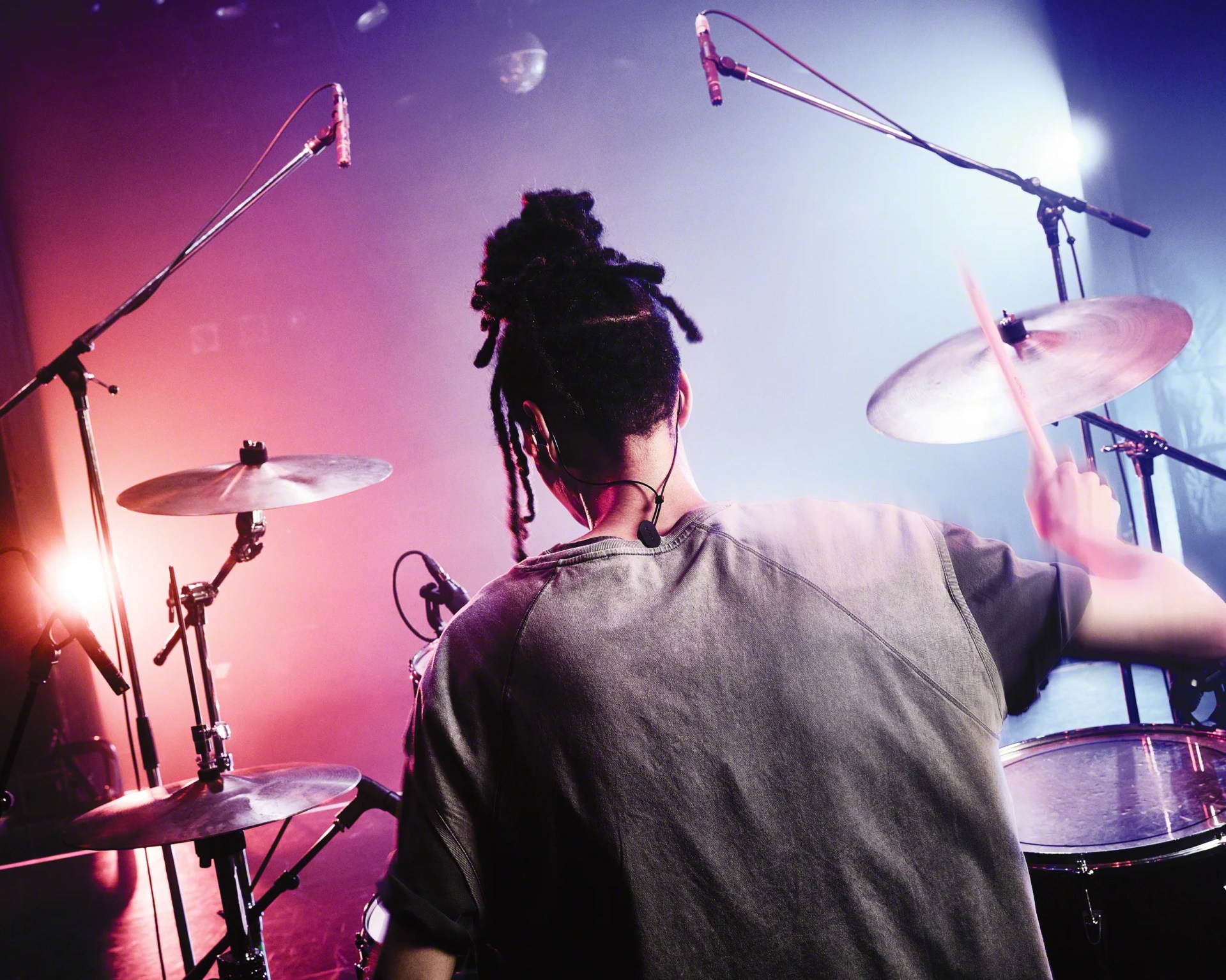 Drummer with dreadlocks playing a drum set under purple and blue stage lights.