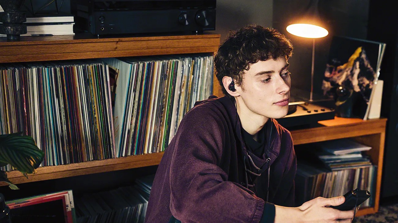 Young person wearing dark earbuds and a purple hoodie, holding a remote, sitting near a wooden shelf filled with vinyl records and a lit table lamp.