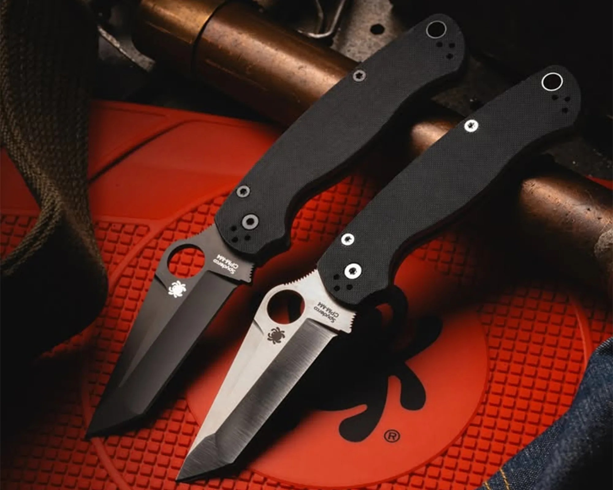 Two Spyderco folding knives with black textured handles and partially serrated blades on an orange textured mat.