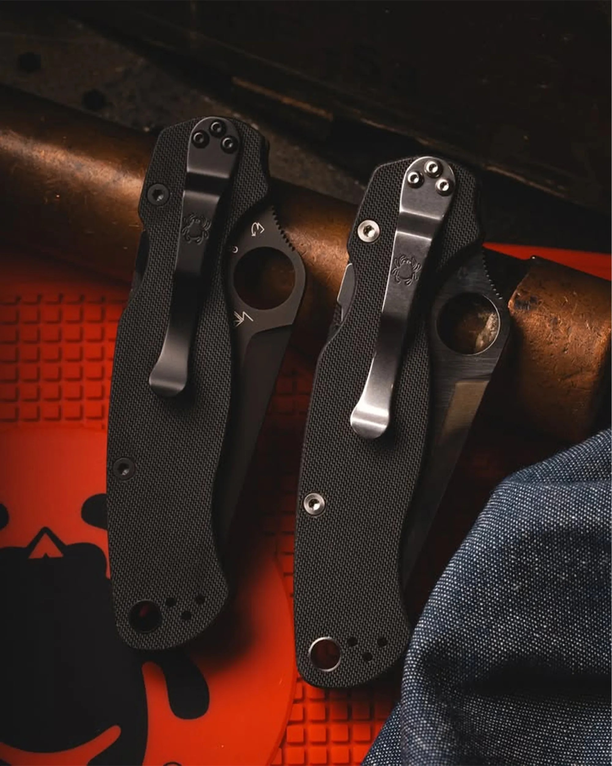 Two black folding knives with textured handles and metal clips resting on a brown cylindrical object and a red surface.