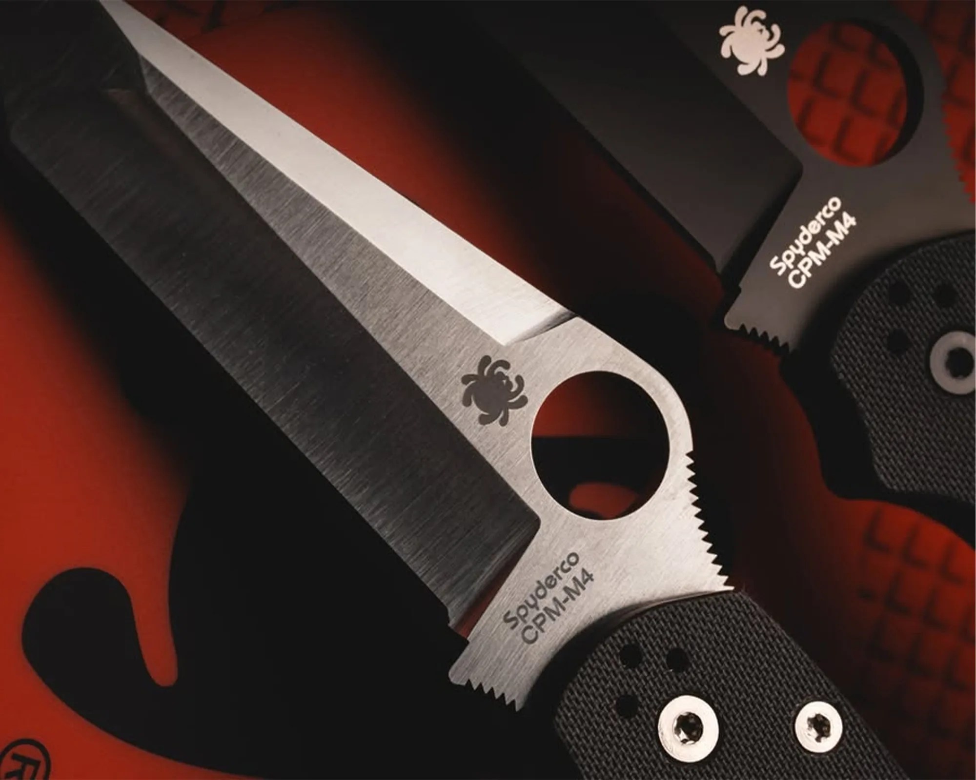 Close-up of two Spyderco CPM-M4 knives with partially serrated stainless steel blades and black textured handles on a red surface.