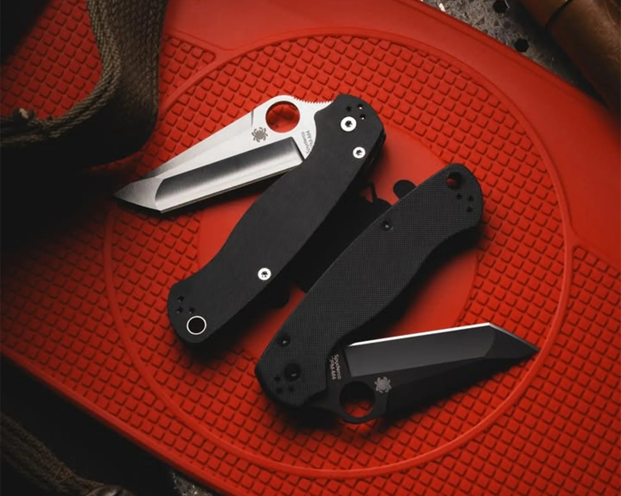 Two black folding knives with partially open blades on a textured red surface.
