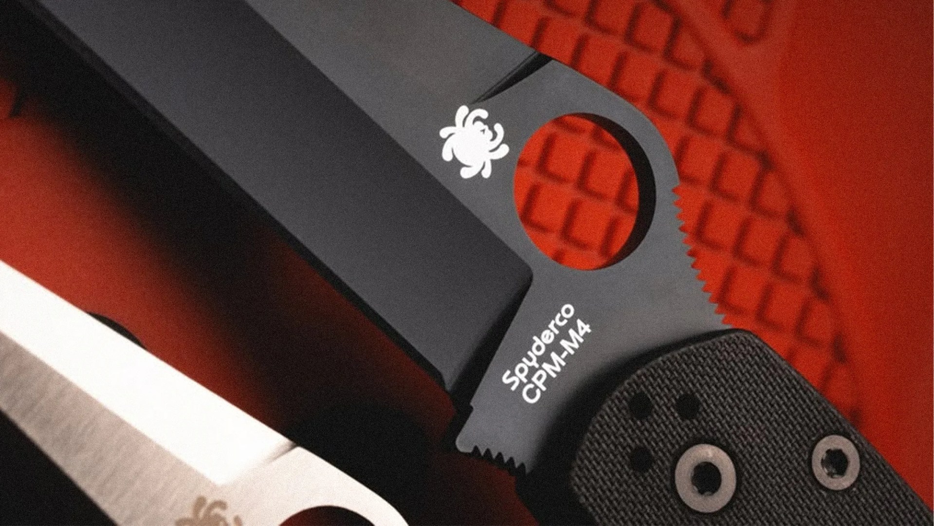 Close-up of a black Spyderco CPM-M4 folding knife blade with a textured handle on a red surface.