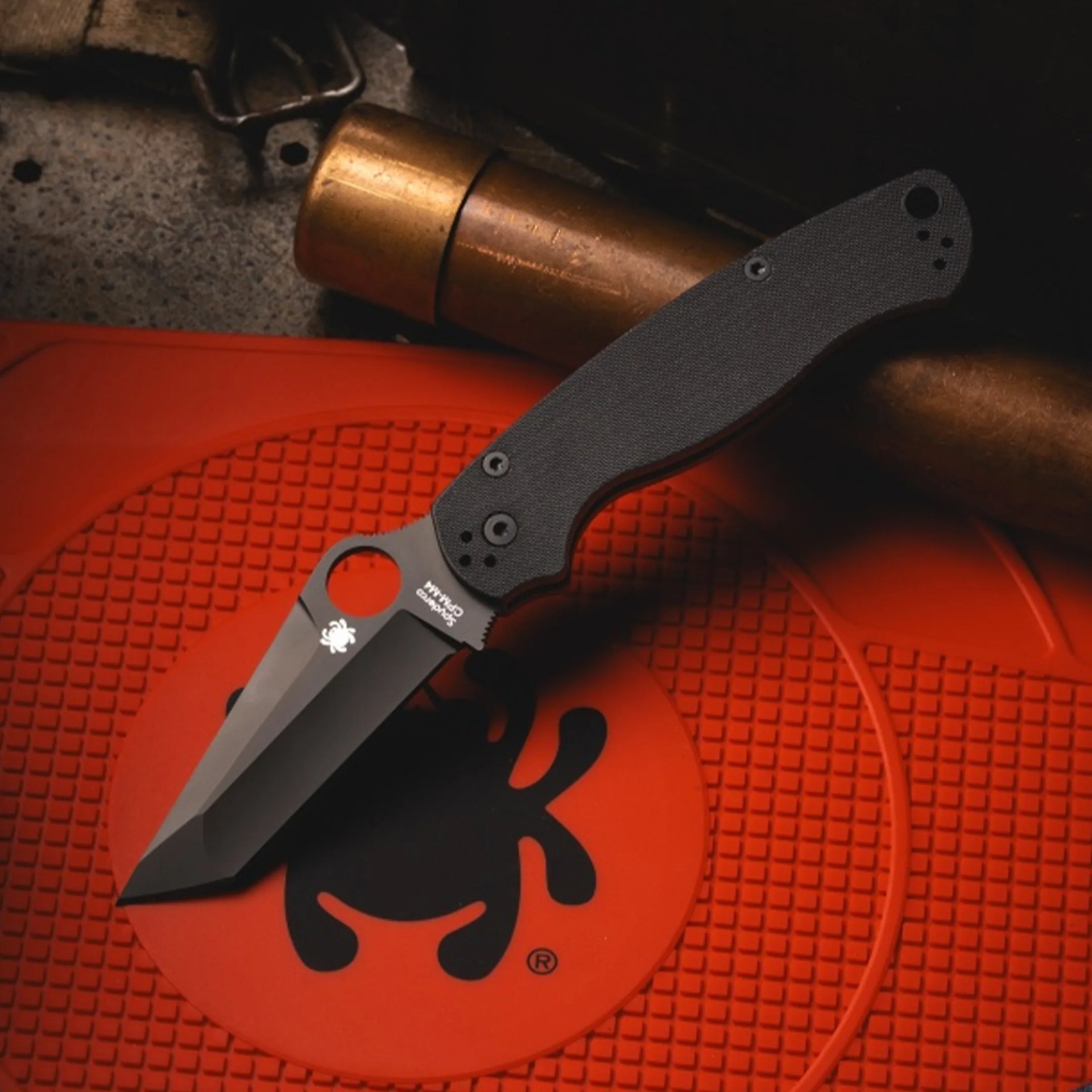 Black folding knife with textured handle on an orange mat featuring a black spider logo.
