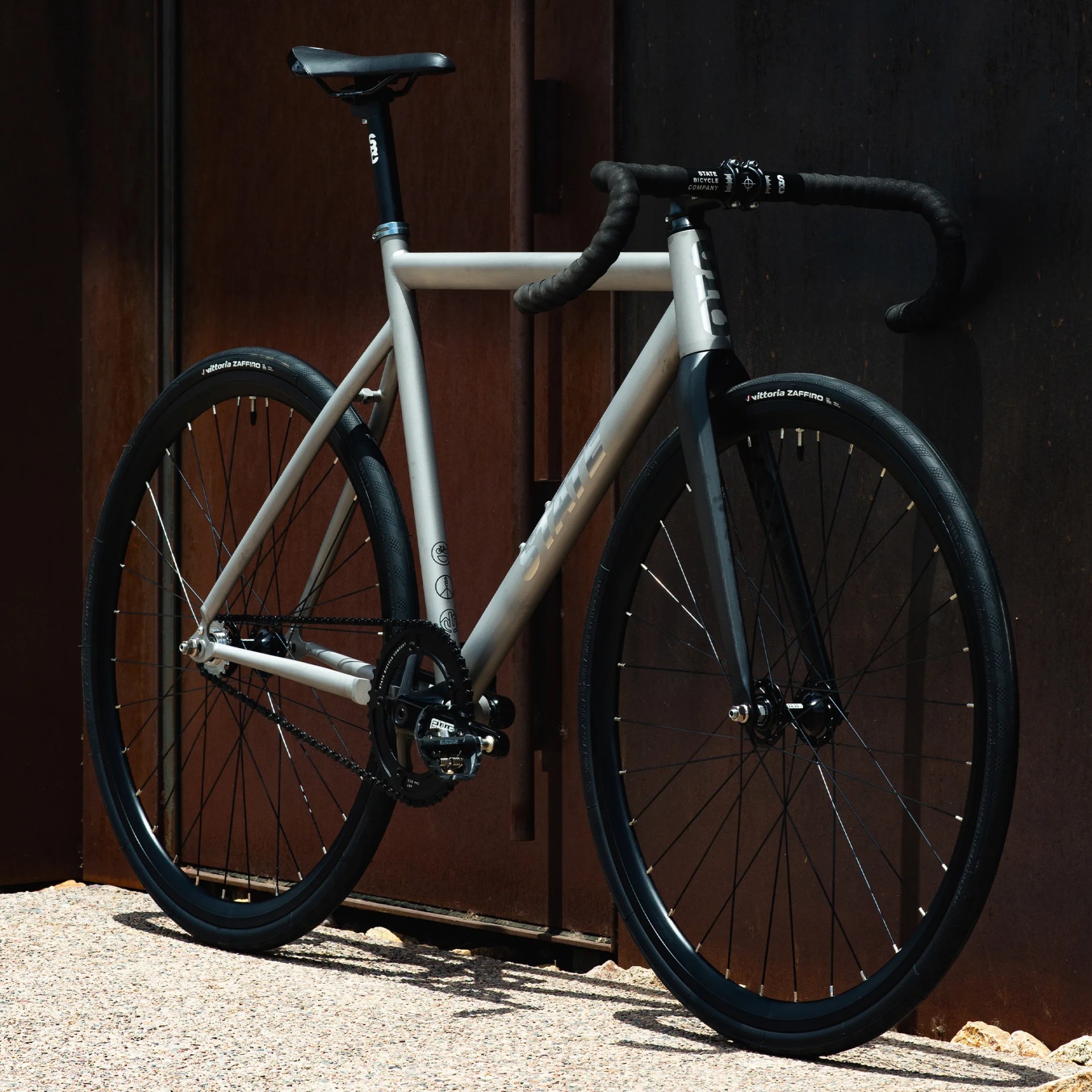 Matte silver and black road bike with drop handlebars and Vittoria Zaffiro tires leaning against a dark metal wall.