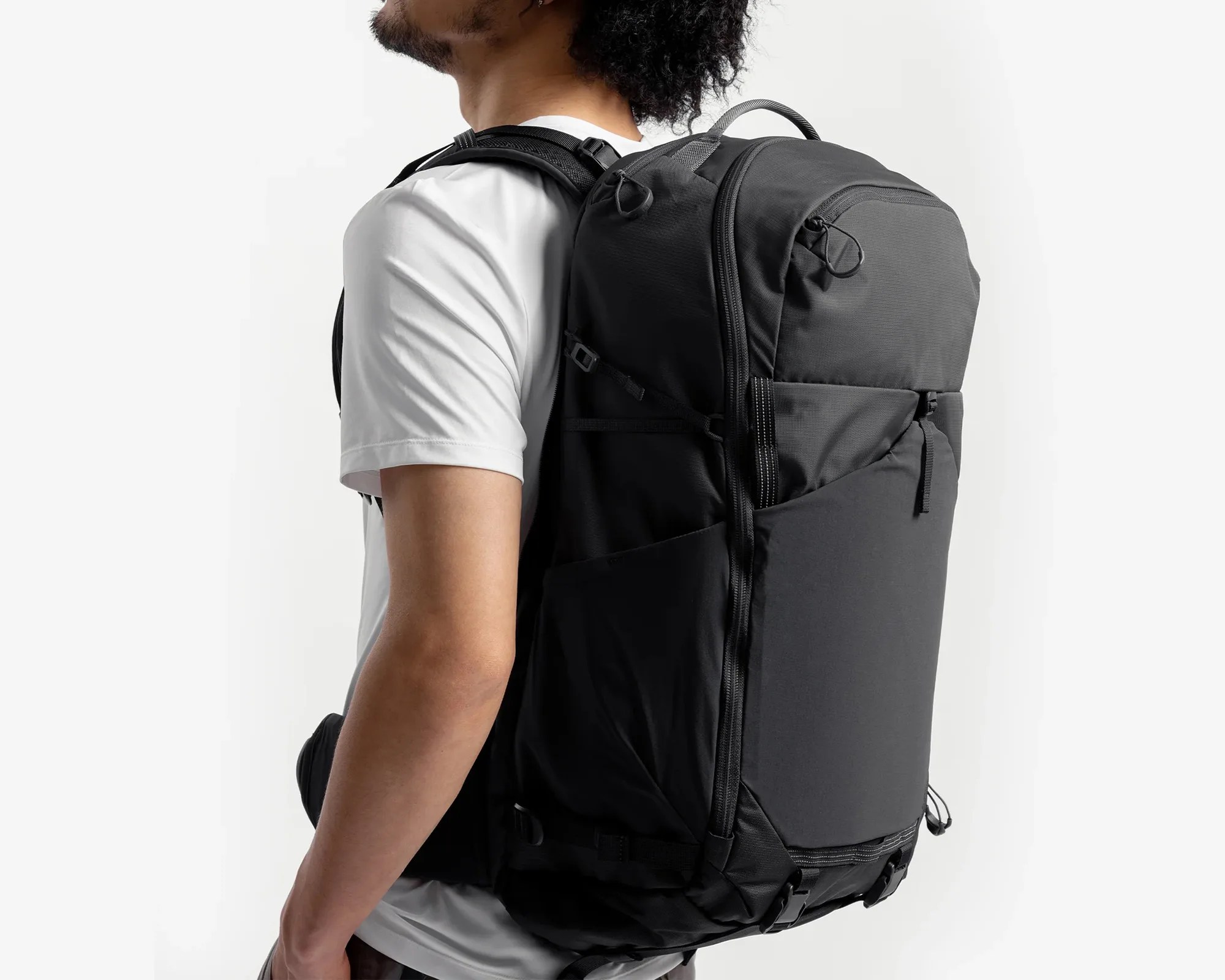Black large backpack with multiple zippers and padded straps worn by a person in a white t-shirt.