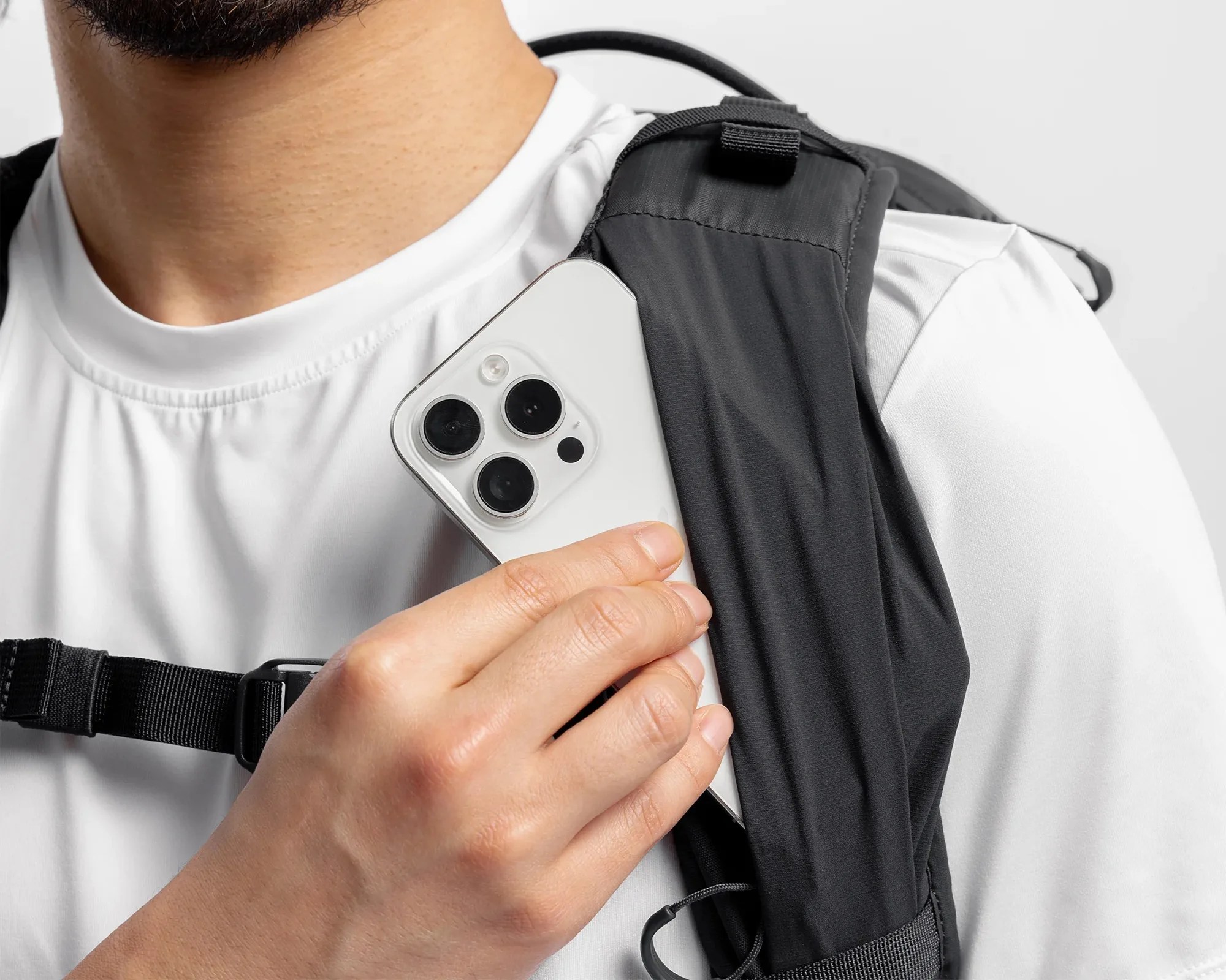 Hand holding a white smartphone with triple camera lenses near a black backpack strap on a person wearing a white shirt.