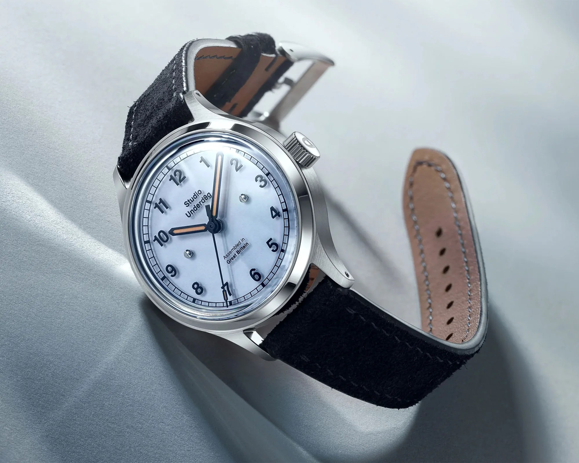 Silver wristwatch with white dial, black numerals, and black suede strap on a light gray surface.