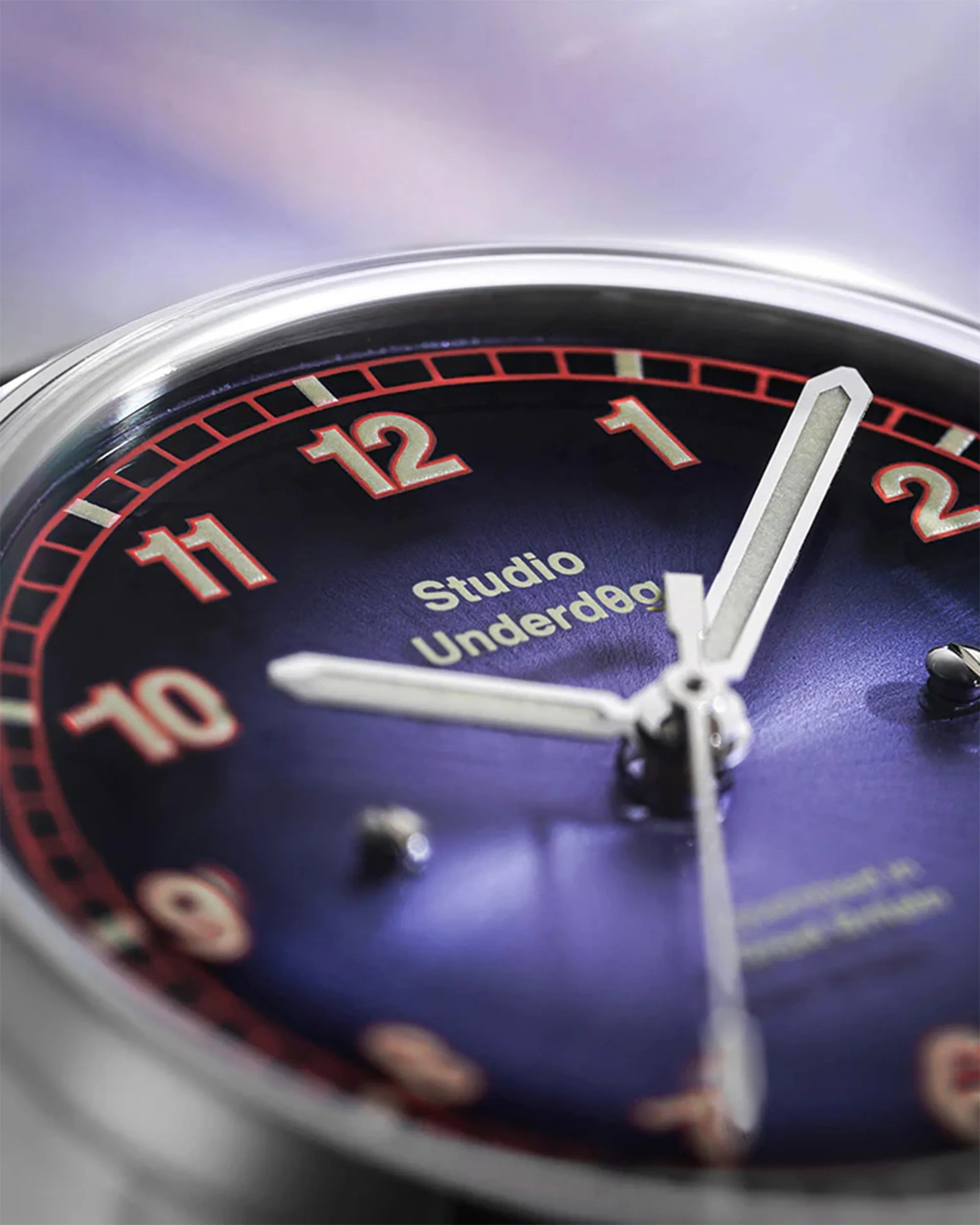 Close-up of a watch face with a blue dial, red-bordered numbers, and silver hands showing the time near 12:05.