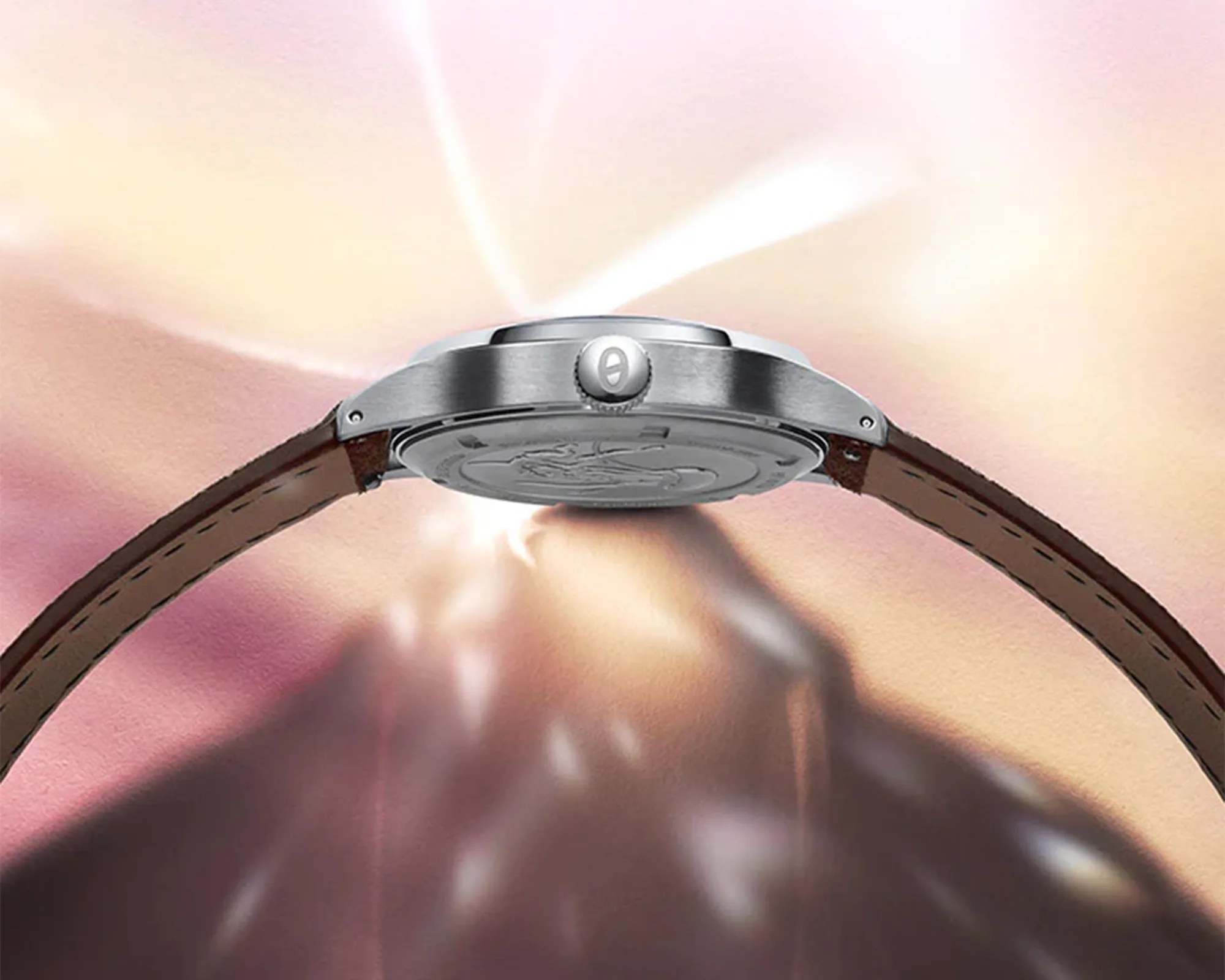 Side view of a silver watch case with a crown and brown leather strap against a blurred pink and beige background.