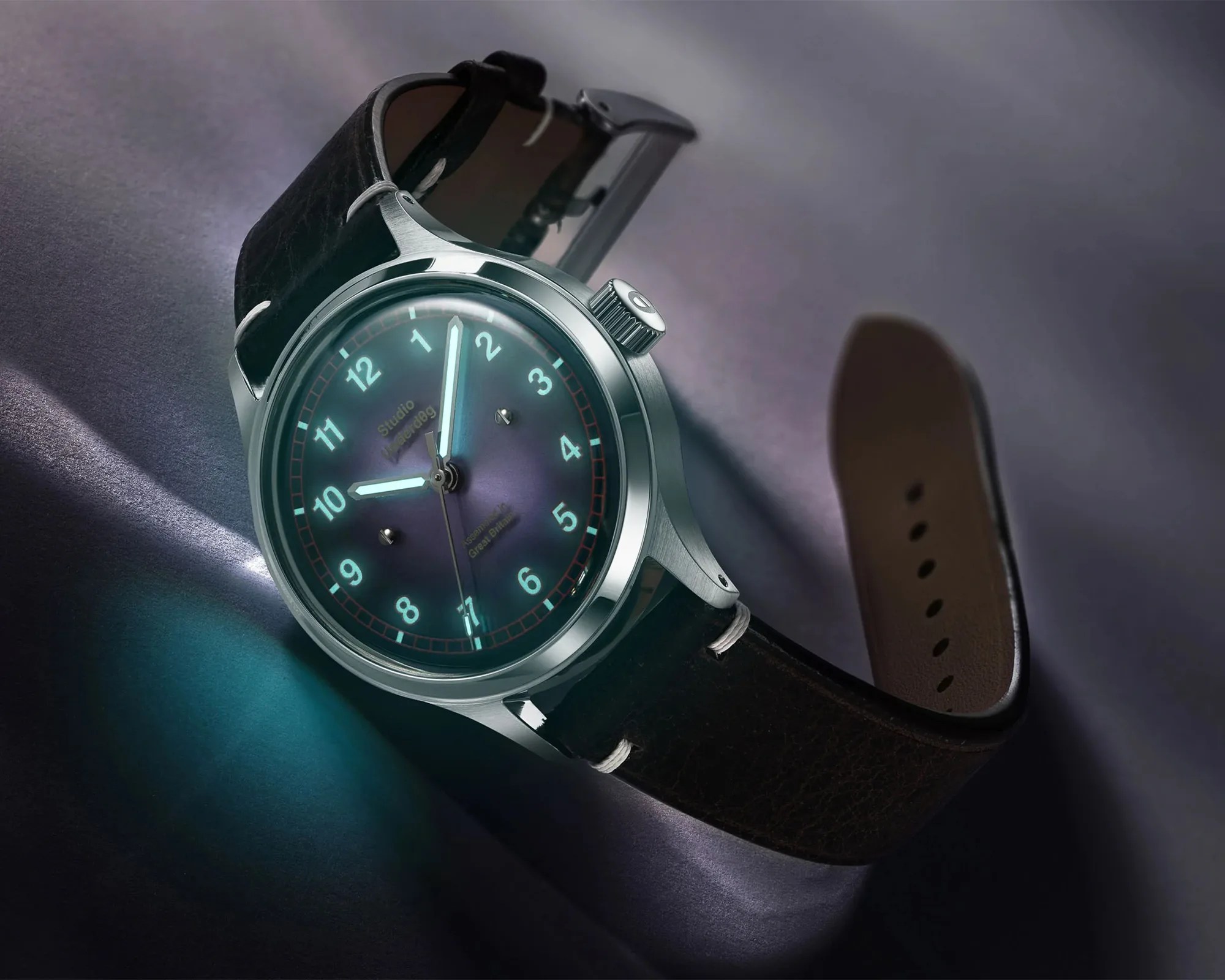 Round silver wristwatch with glowing blue dial and black leather strap on a dark surface.