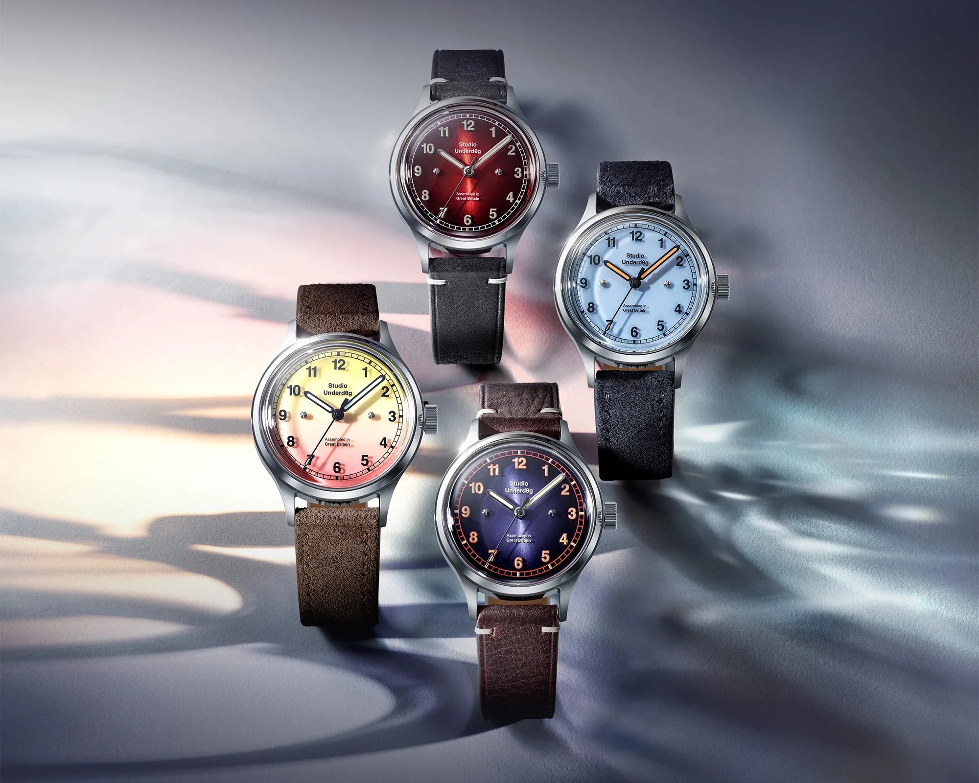 Four Studio Underd0g wristwatches with leather straps in red, blue, yellow, and purple dials on a gray surface.
