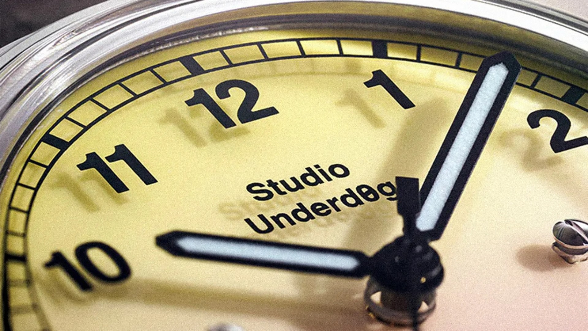 Close-up of a yellow clock face showing numbers 10 to 2 with black hour and minute hands and "Studio Underdog" text.