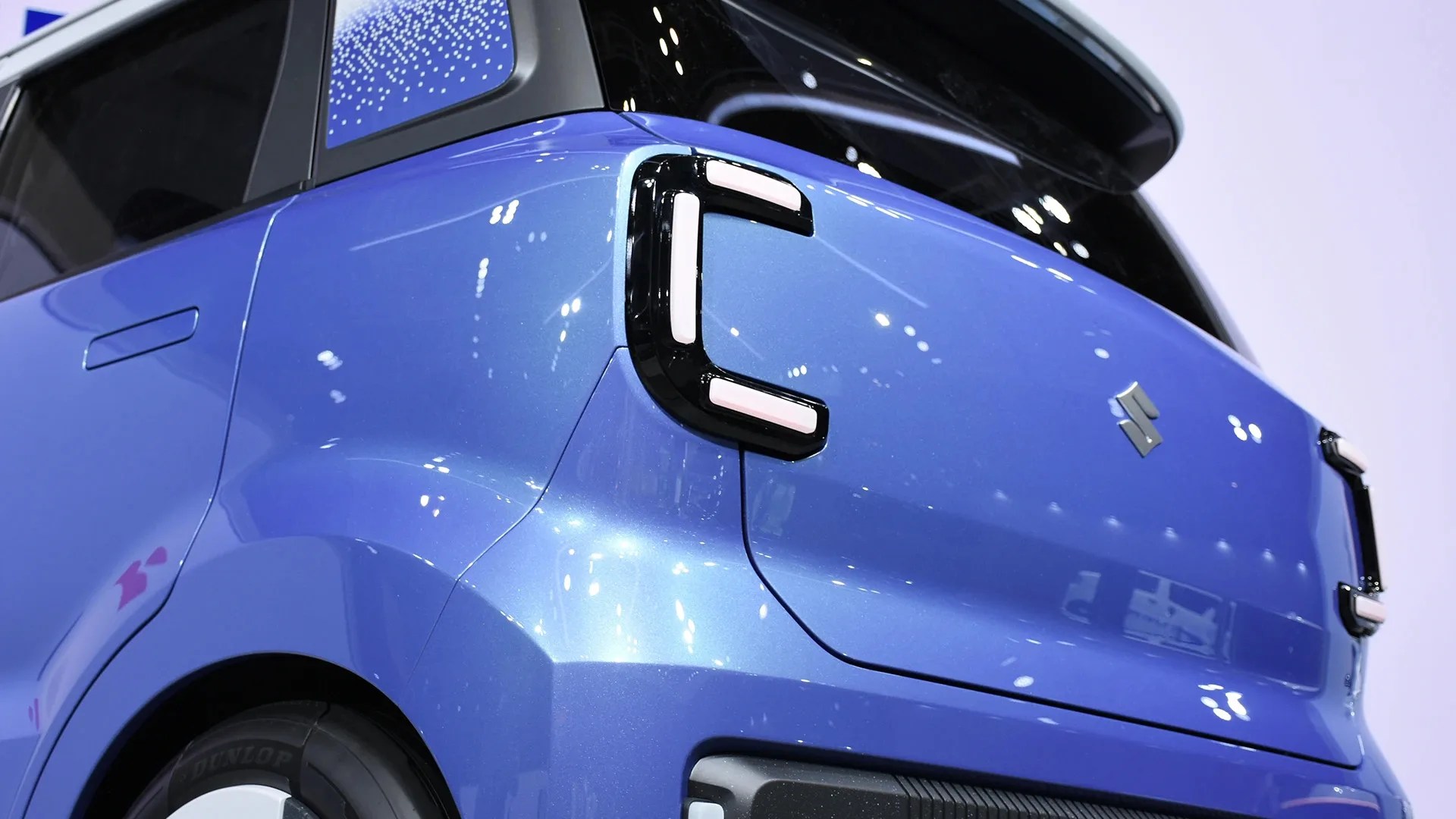 Rear corner of a blue Suzuki car with distinctive C-shaped taillights and black roof spoiler.