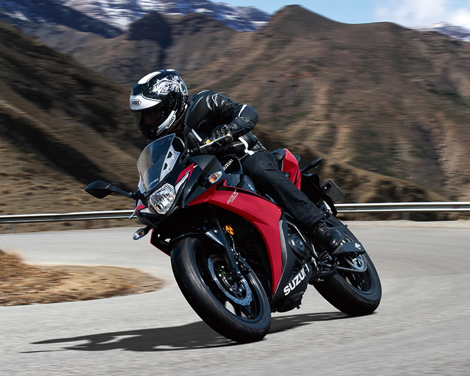 Person wearing black riding gear and white helmet leaning into a turn on a red and black Suzuki motorcycle on a mountain road.