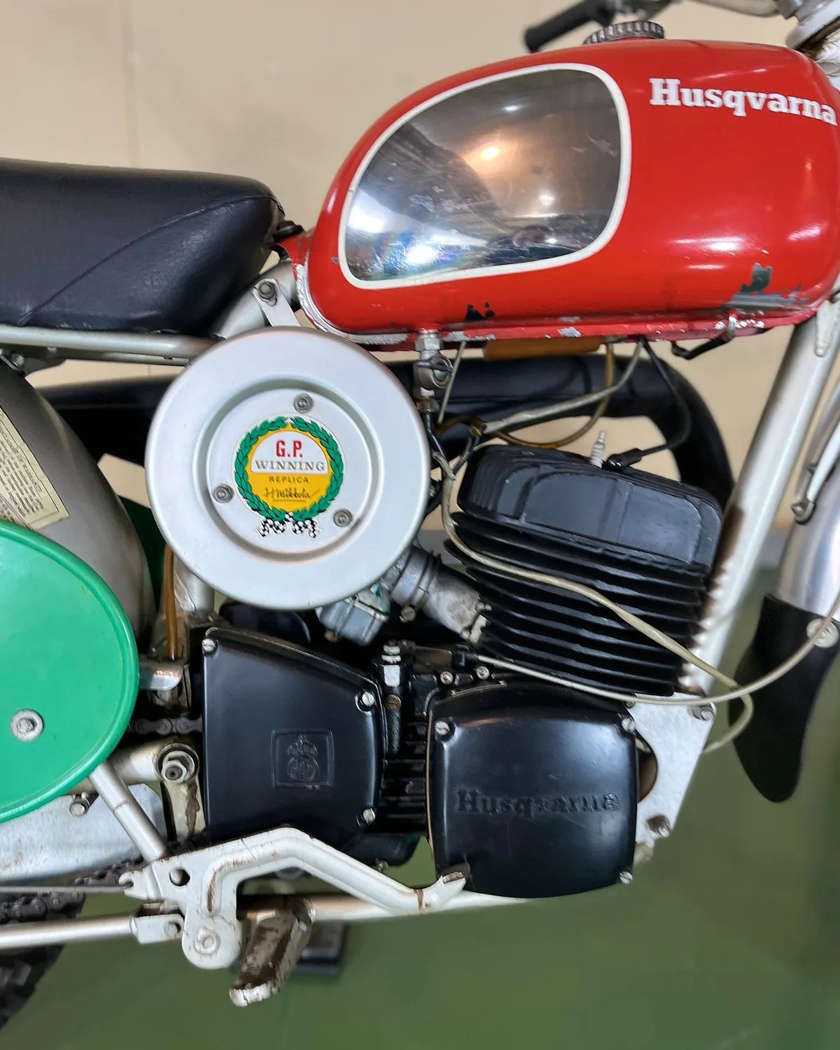 Close-up of a vintage Husqvarna motorcycle engine with a red fuel tank and a circular silver air filter cover labeled "G.P. Winning Replica.