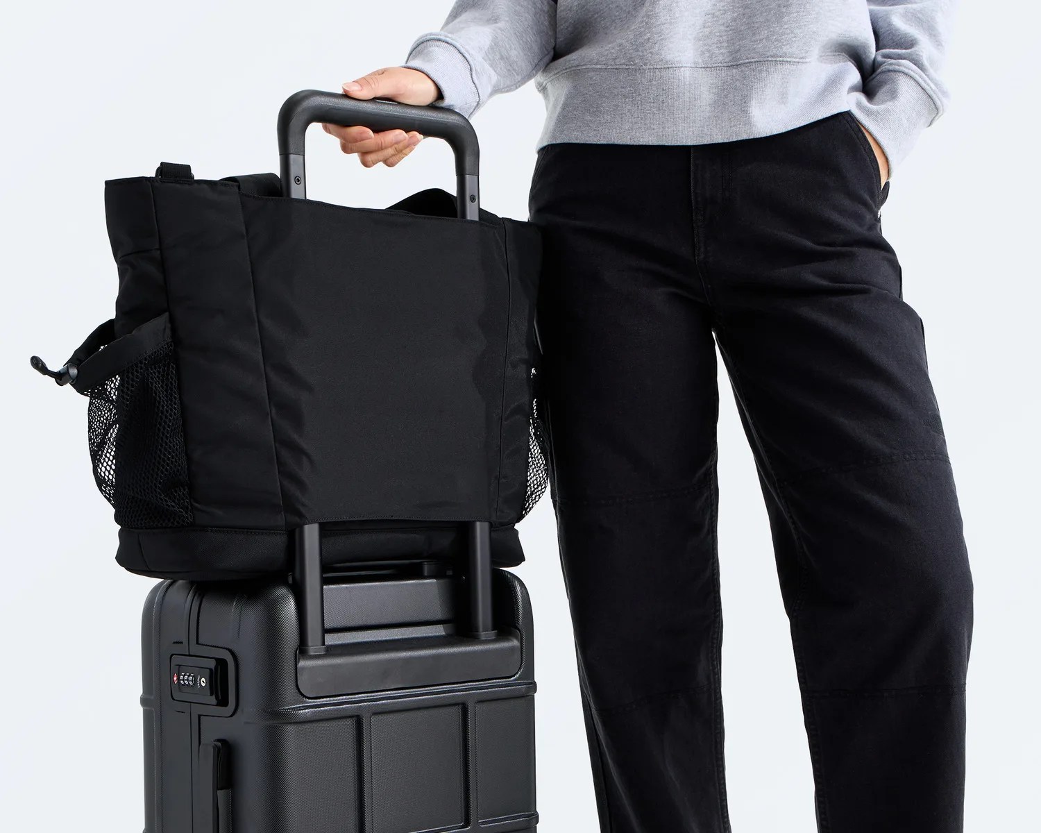 Black soft-sided tote bag with mesh side pockets attached to the handle of a black hard-shell suitcase.