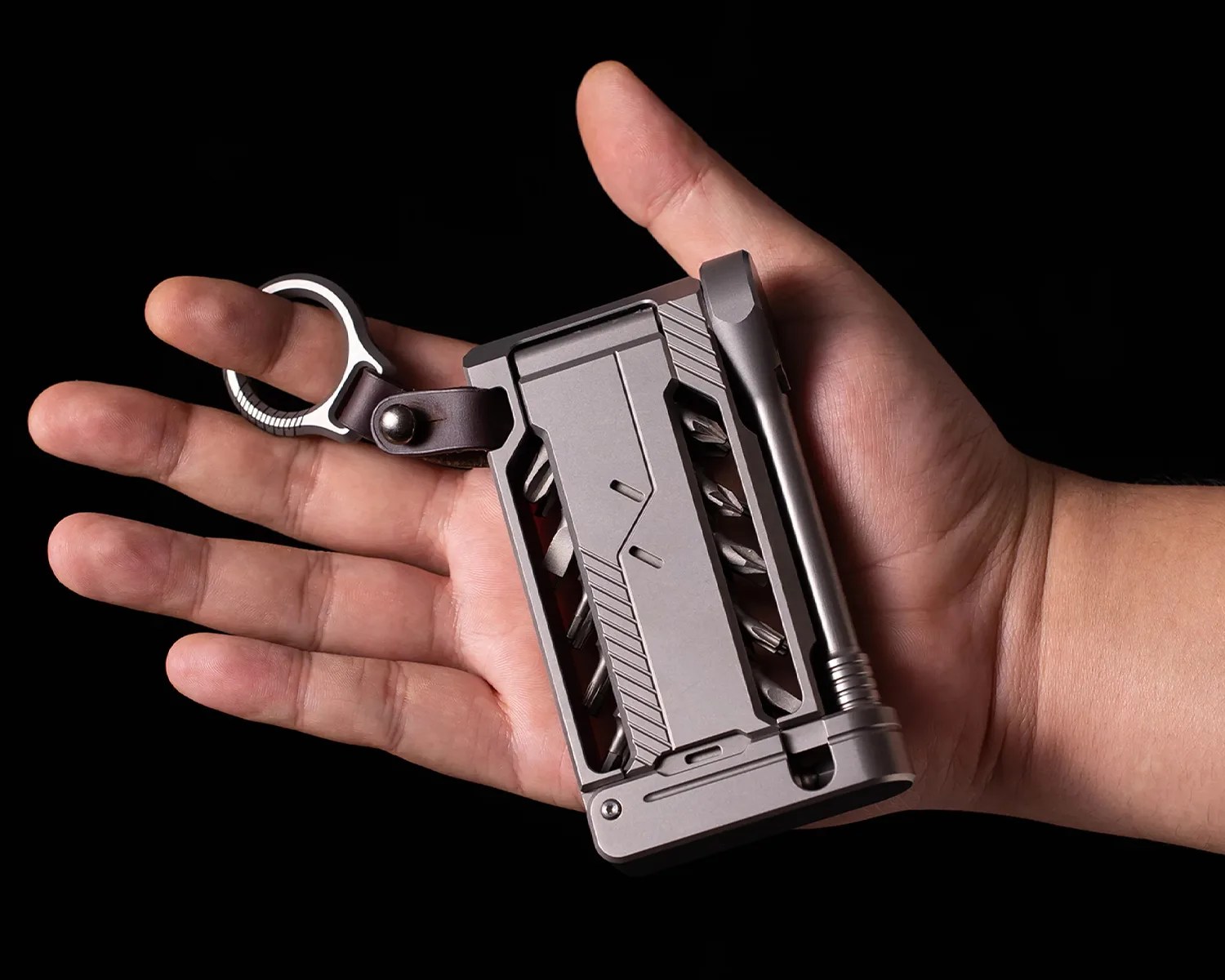 Compact gray multi-tool with screwdriver bits and a keyring held in an open hand against a black background.