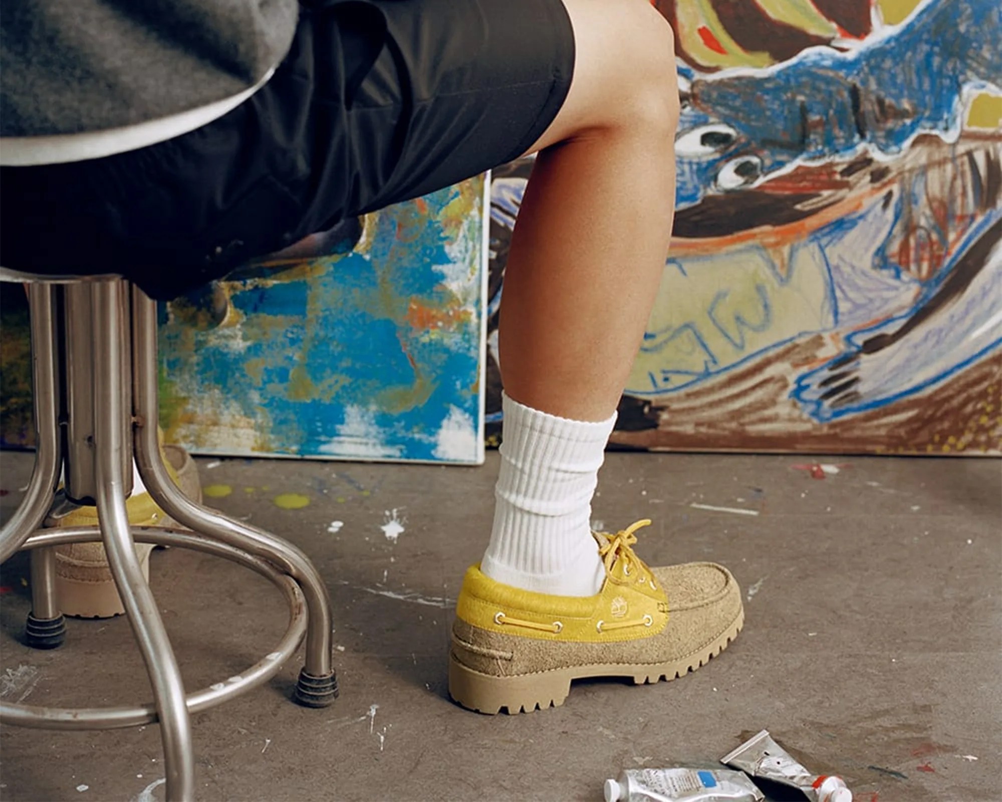 Person wearing yellow and beige suede shoes with white ribbed socks and black shorts sitting on a metal stool in an art studio.