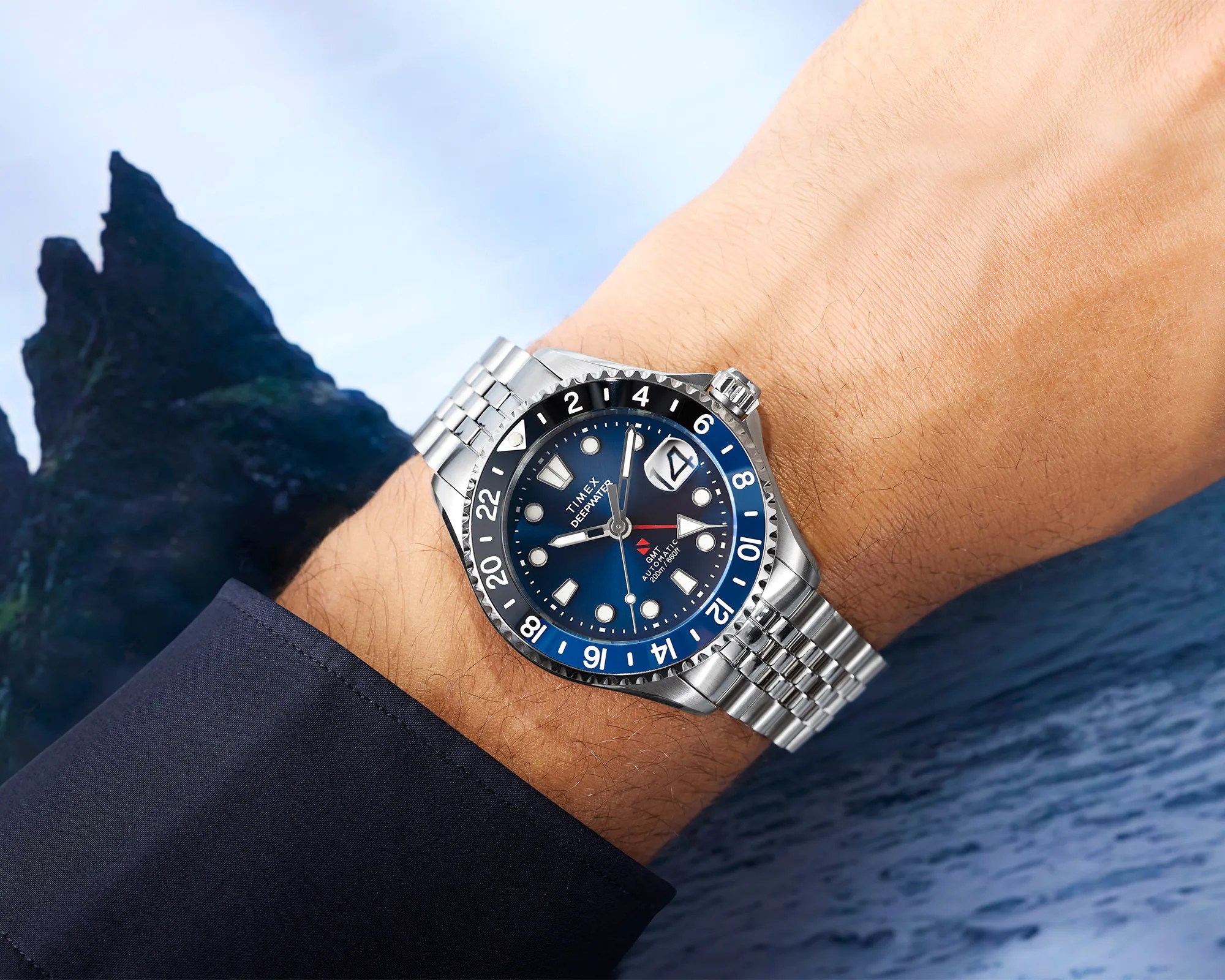 Silver Timex Deepwater GMT automatic watch with blue and black bezel on a wrist over a blue ocean background.