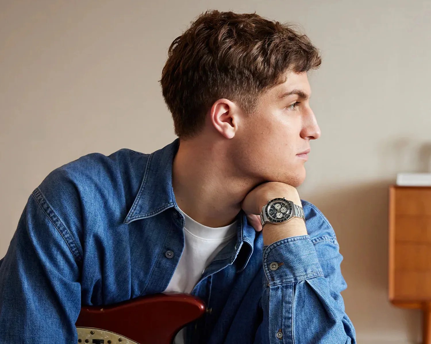 Young man in a blue denim shirt resting his chin on his hand, wearing a silver wristwatch with a black dial.