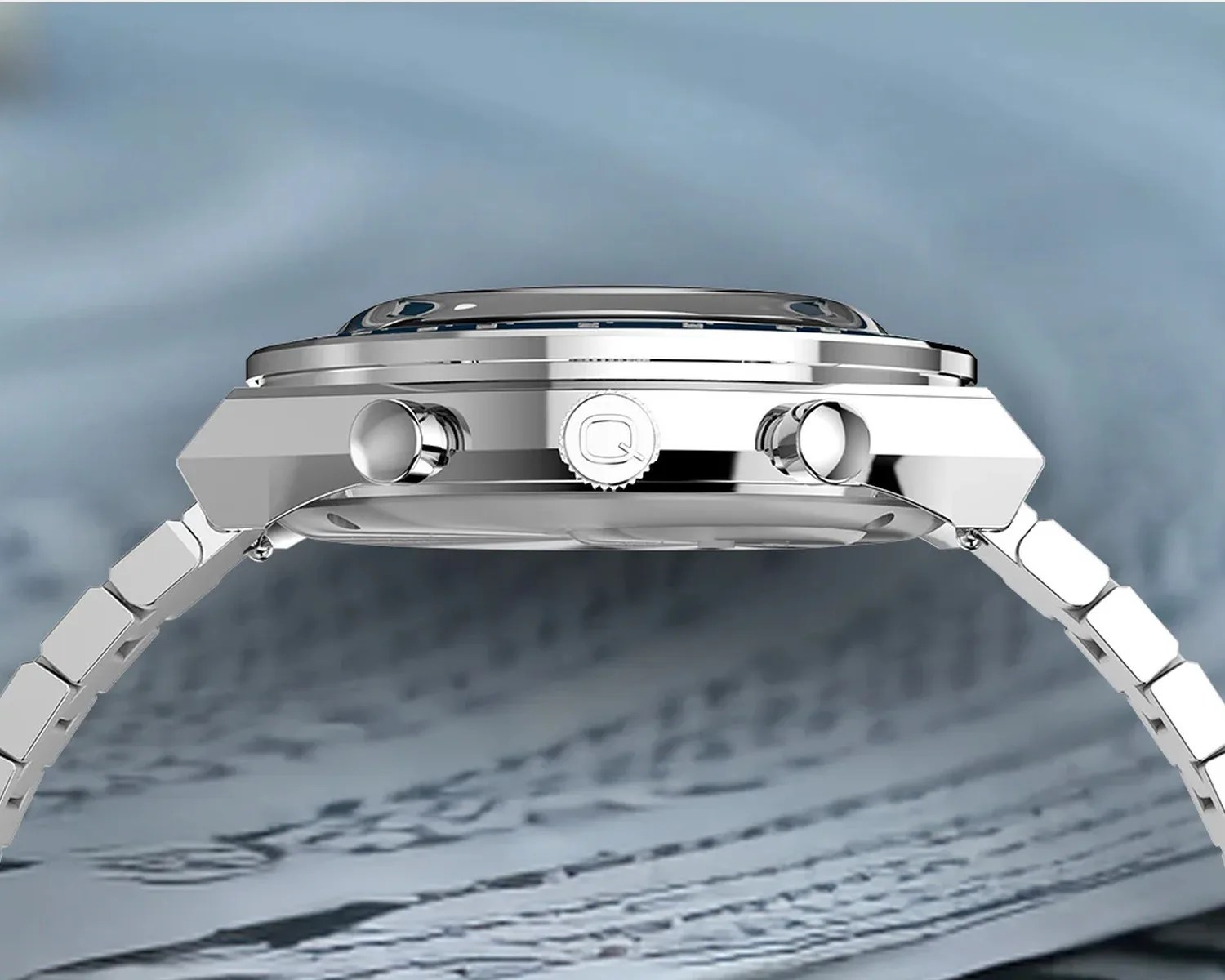 Side view of a silver metal wristwatch with two push buttons and a crown engraved with a "Q" on a blurred blue background.