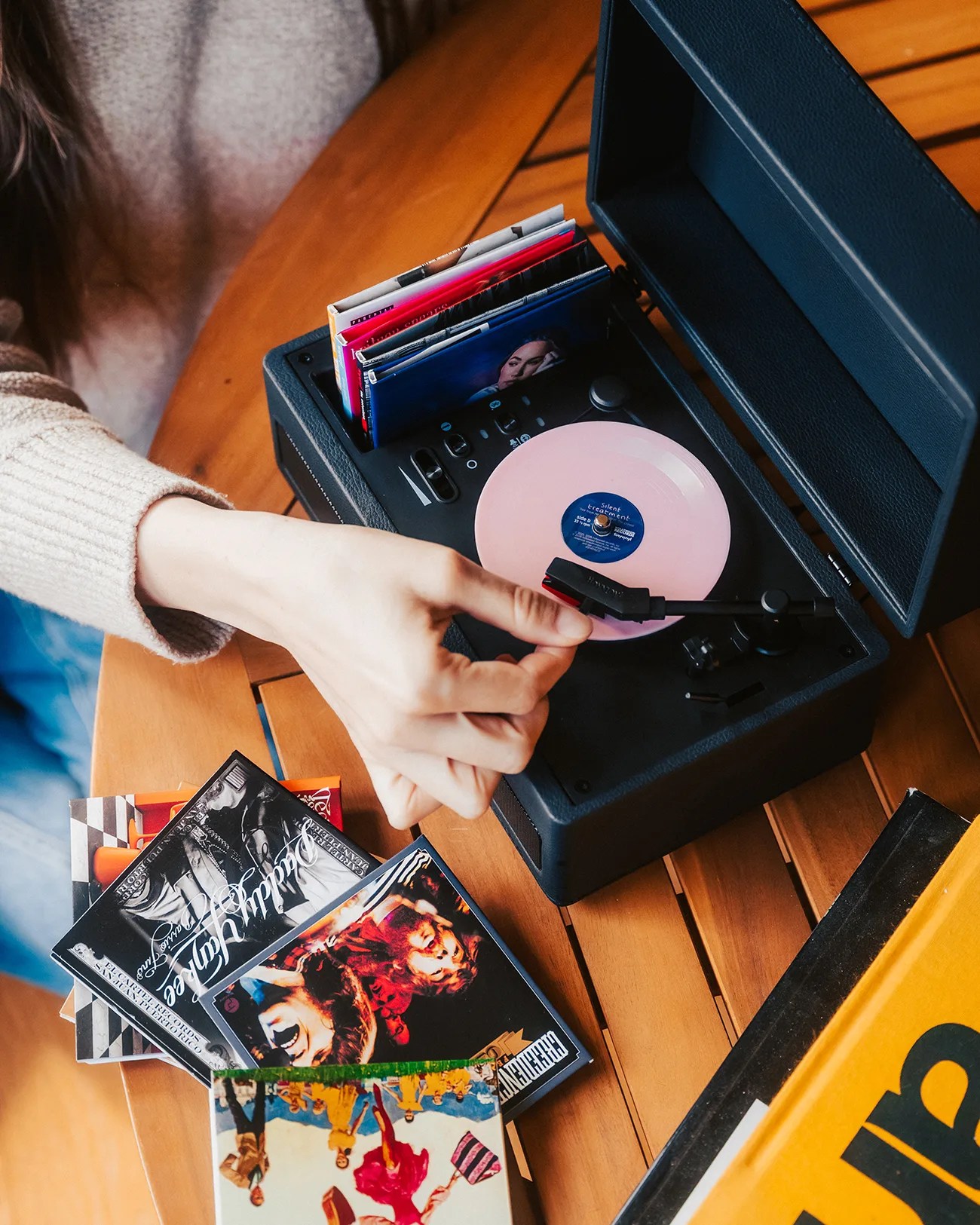 Hand adjusting the tonearm of a black portable record player with a pink vinyl on a wooden table.