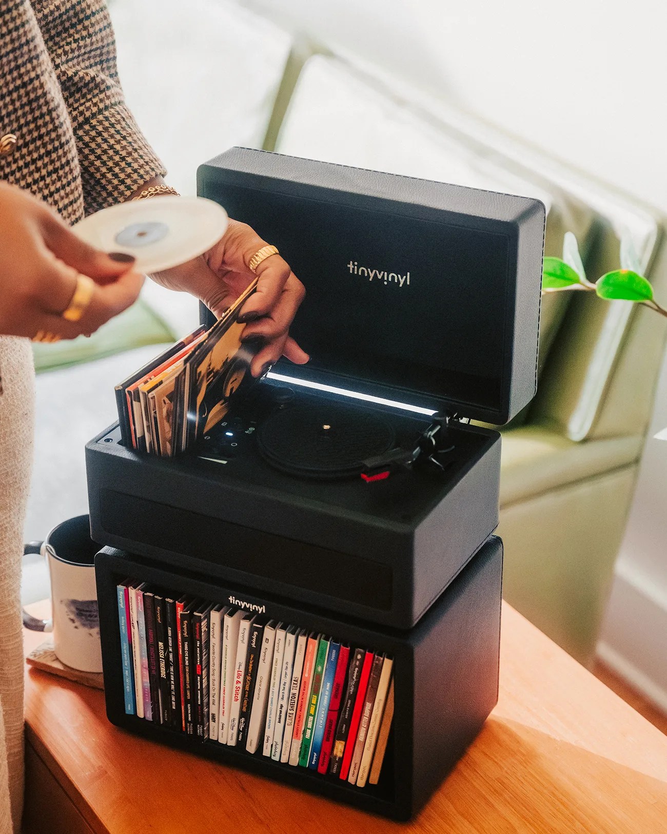 Black TinyVinyl record player with open lid and a person holding a vinyl record and album covers on a wooden table.