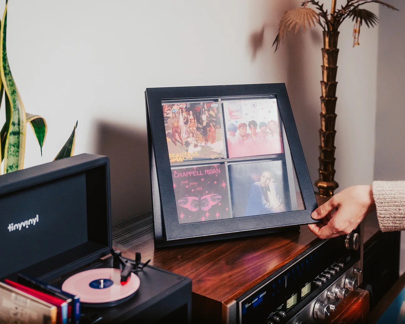 Black square frame displaying four vinyl record covers on a wooden stereo cabinet with a hand adjusting it.